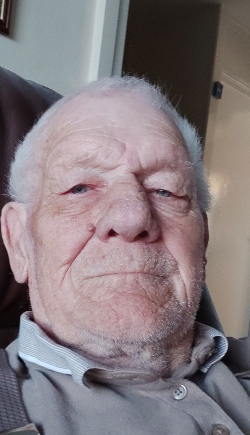 Elderly man with short white hair and stubble, wearing a collared shirt, looking directly at the camera with a neutral expression.