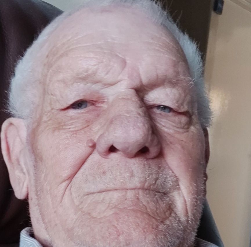 Elderly man with short white hair and stubble, wearing a collared shirt, looking directly at the camera with a neutral expression.
