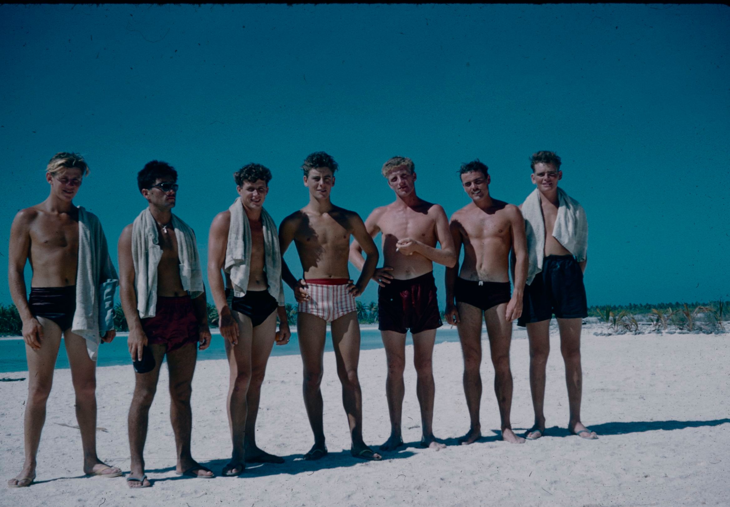 Seven young men pose in swimsuits on a white beach of Christmas Island. Four of the men have towels draped around their necks or off their shoulders.