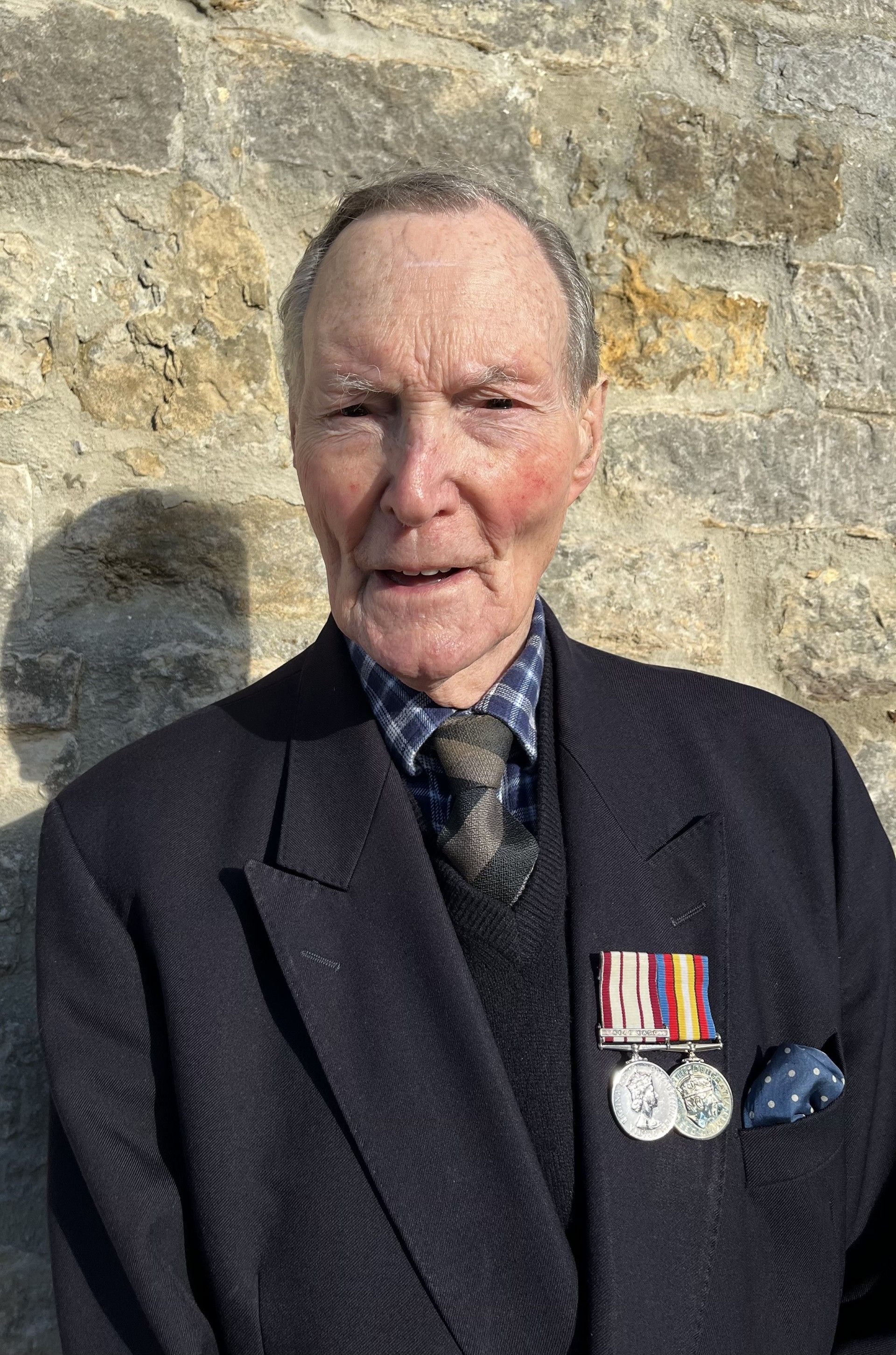Elderly man with short hair wearing a dark blazer, checkered shirt, and tie, with several military medals pinned to his chest, standing against a stone wall.