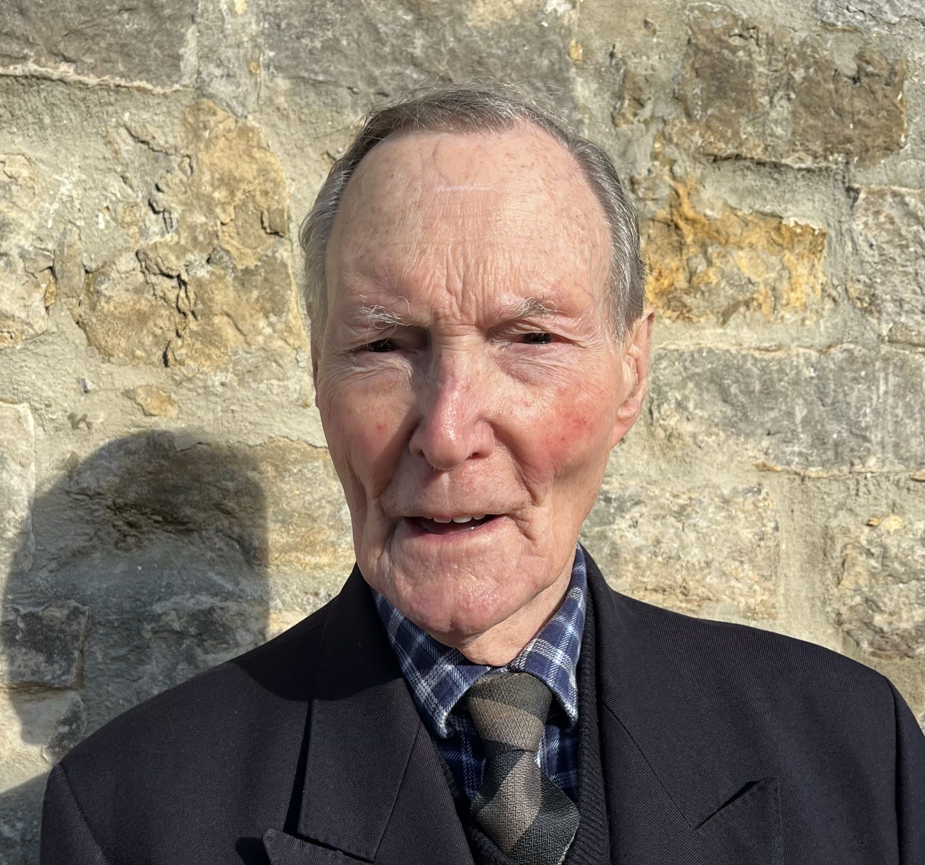 Elderly man with short hair wearing a dark blazer, checkered shirt, and tie, with several military medals pinned to his chest, standing against a stone wall.