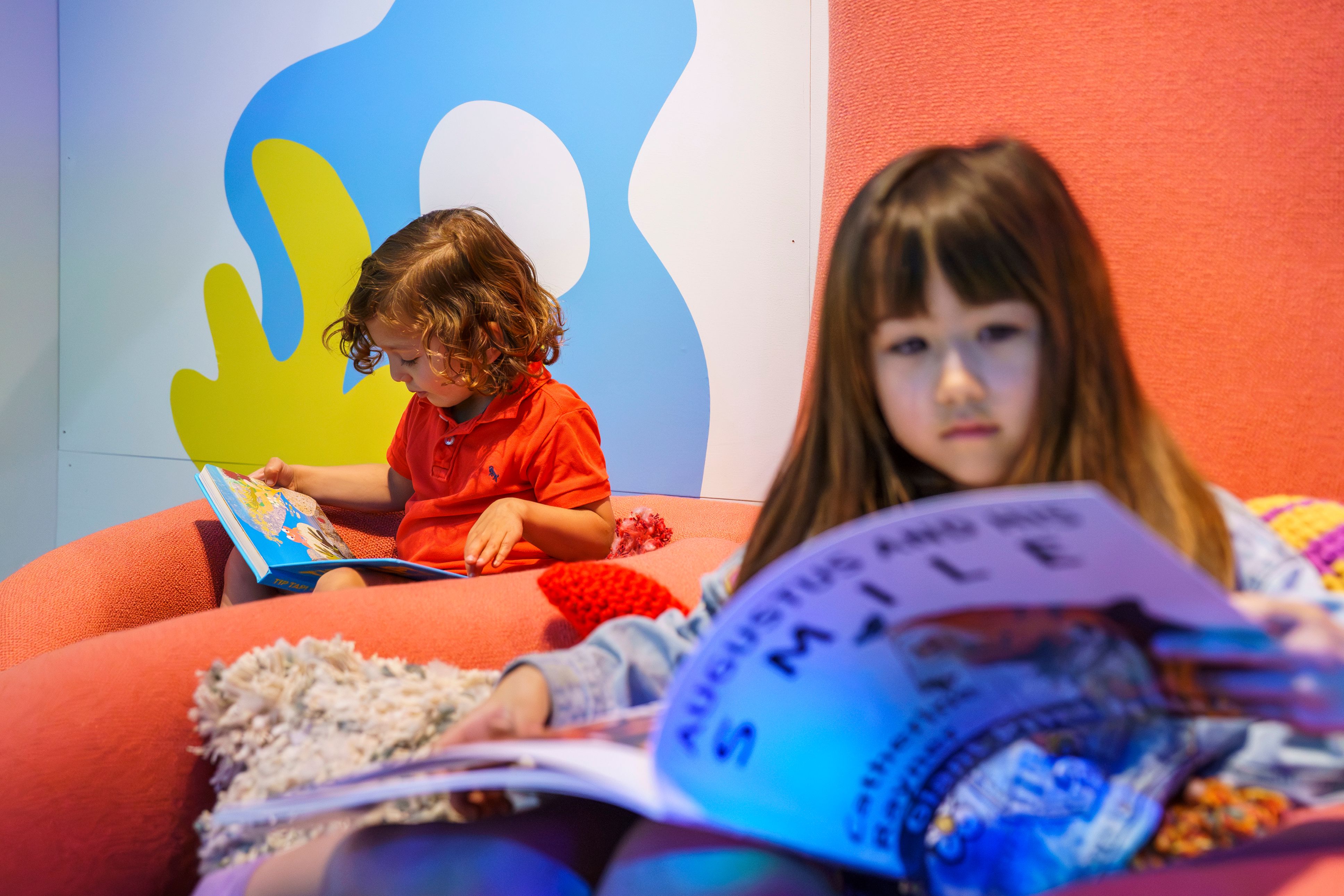 Two children sit on bright, soft seats, reading books in the British Library learning area.