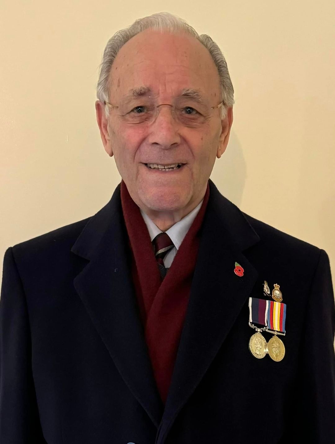 Elderly man smiling, wearing glasses, a dark coat with a red scarf, and a tie, with several military medals and a poppy pin displayed on his chest.
