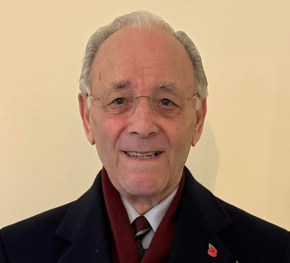 Elderly man smiling, wearing glasses, a dark coat with a red scarf, and a tie, with several military medals and a poppy pin displayed on his chest.