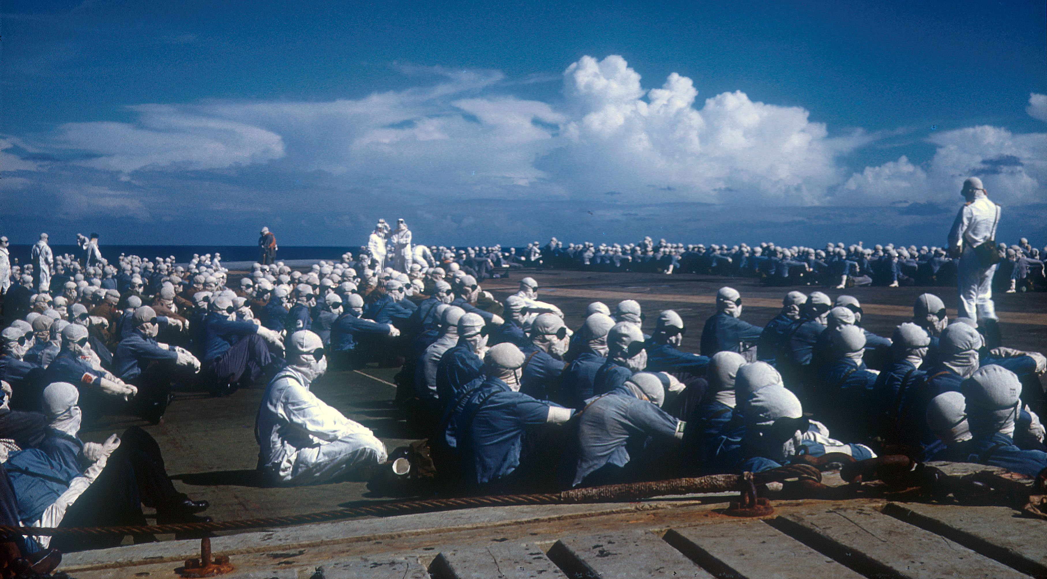 Naval and scientific personnel wearing anti-flash gear, sitting on the floor and all looking in the same direction. A few men are standing up.