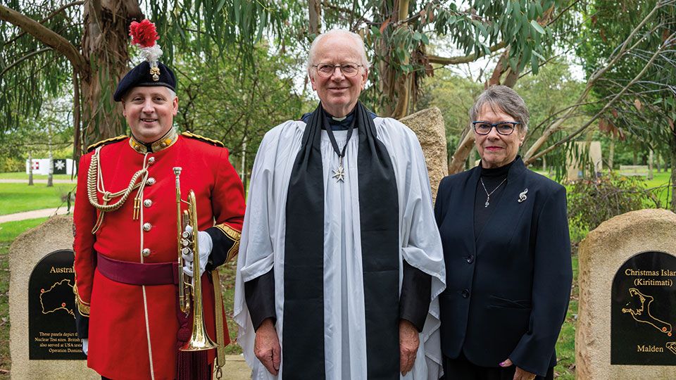 The Very Reverend Nicholas Frayling stands in front of the NMA memorial whilst wearing a white vestment with black scarf.