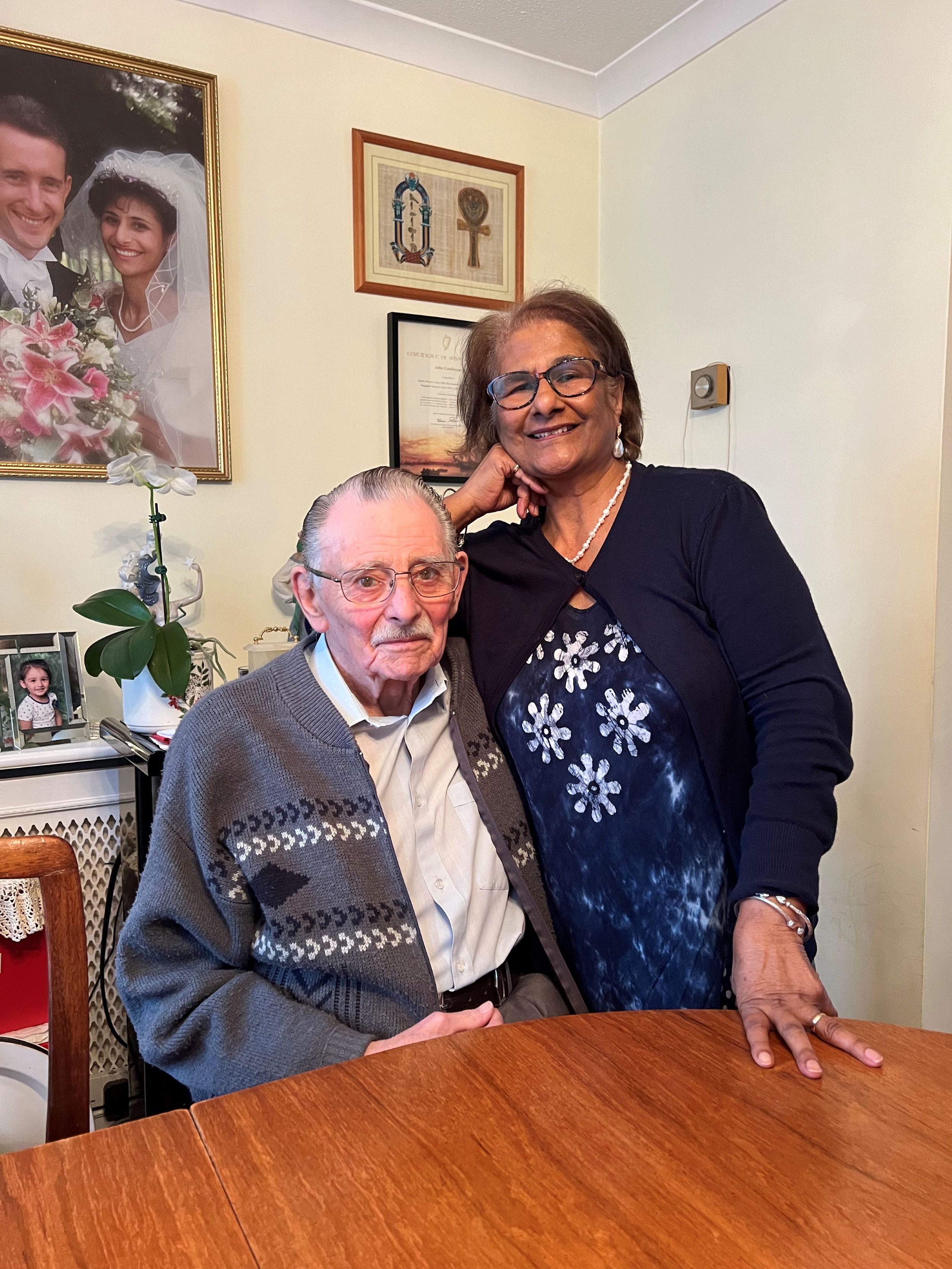 Brian Tomlinson sits at a table in the dining room of his family home, accompanied by his wife, who stands behind him with her hand on his shoulder.