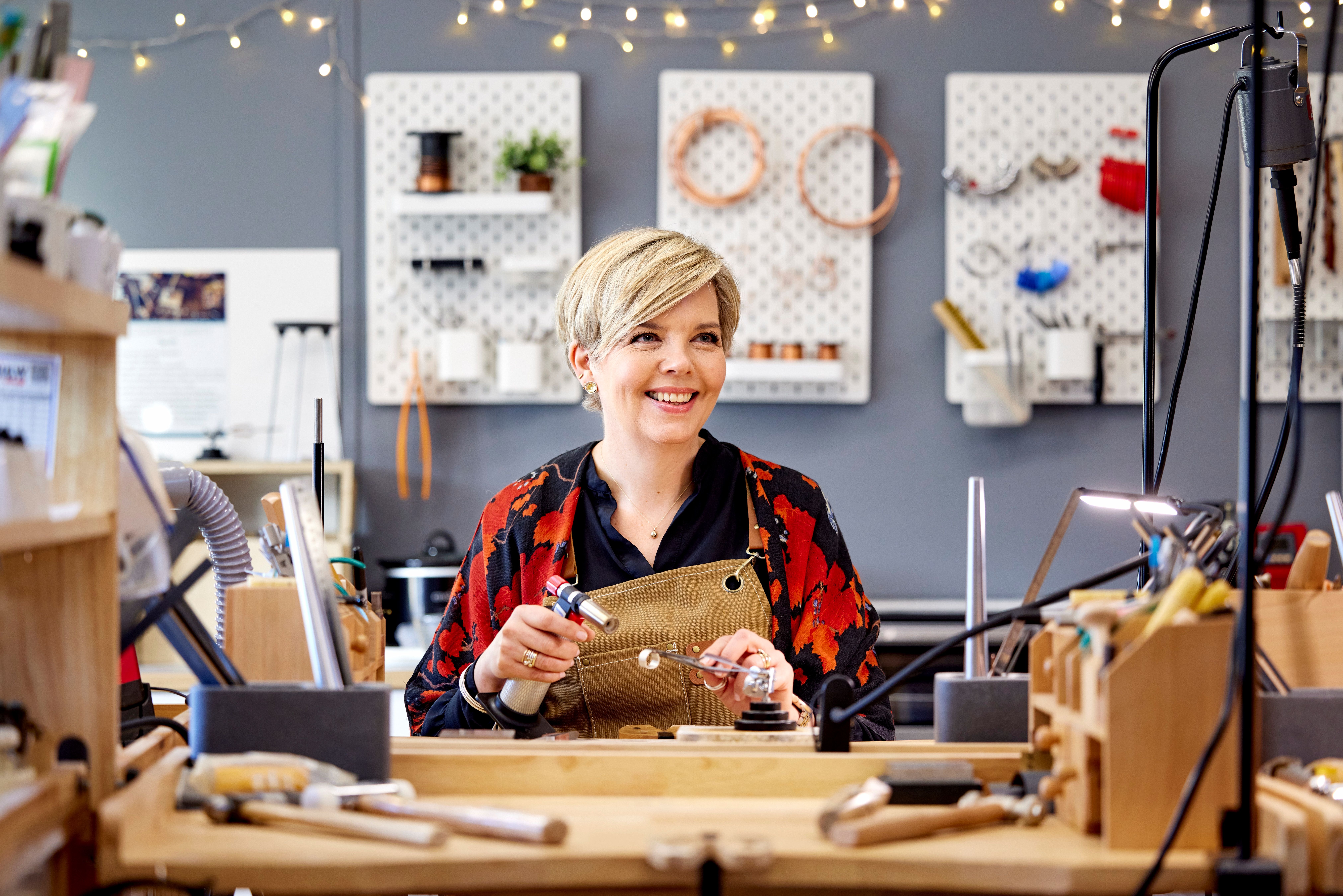 A woman sites at a workbench, with a small blow torch, doing metal work.
