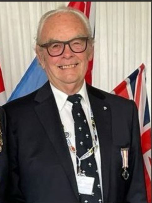 Elderly man smiling, wearing glasses, a dark suit with a patterned tie, a medal on his jacket, and a lanyard with a badge, standing in front of several flags.