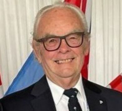 Elderly man smiling, wearing glasses, a dark suit with a patterned tie, a medal on his jacket, and a lanyard with a badge, standing in front of several flags.