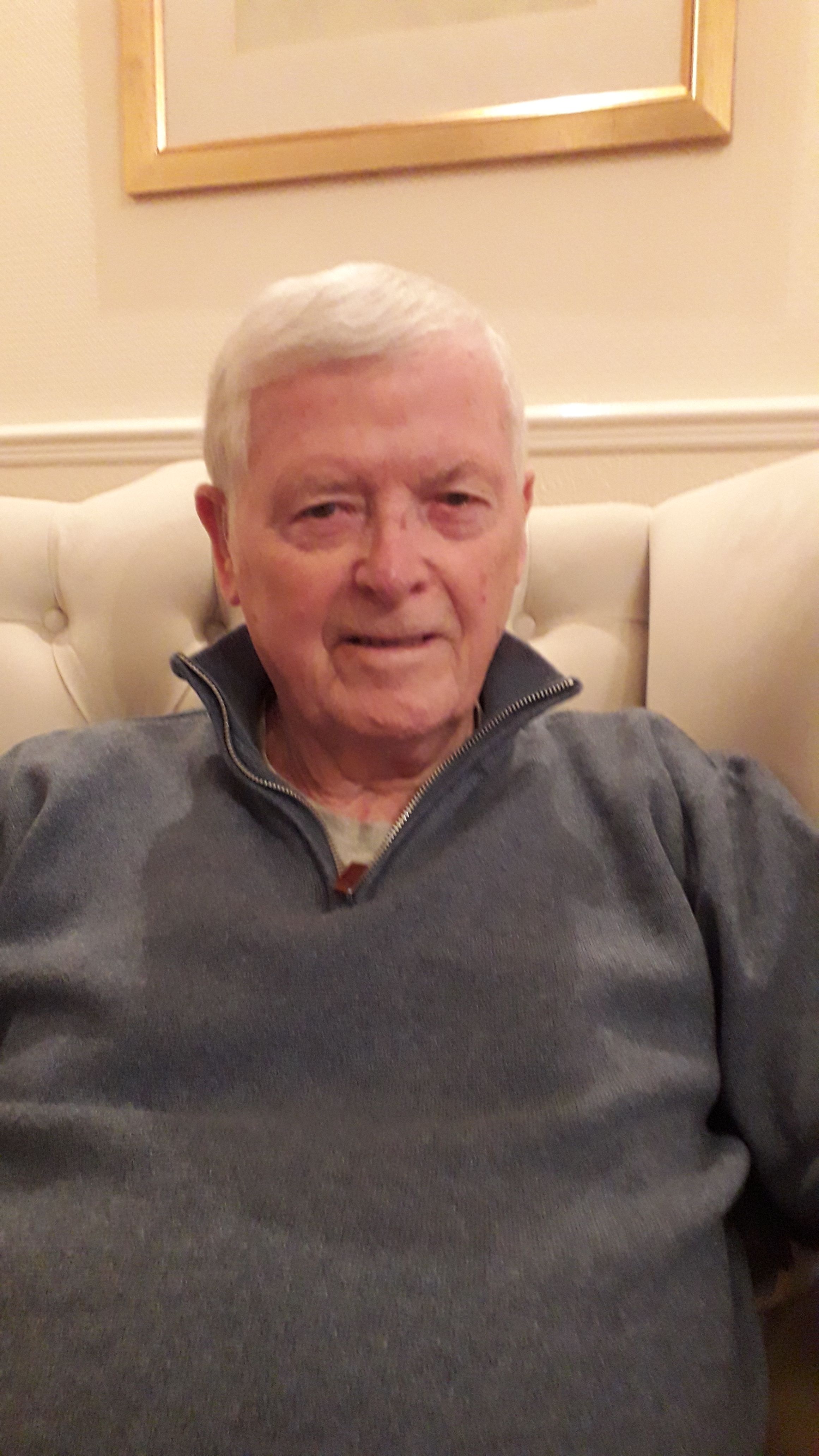 Elderly man with short white hair, smiling gently while seated on a cream upholstered chair, wearing a blue pullover.