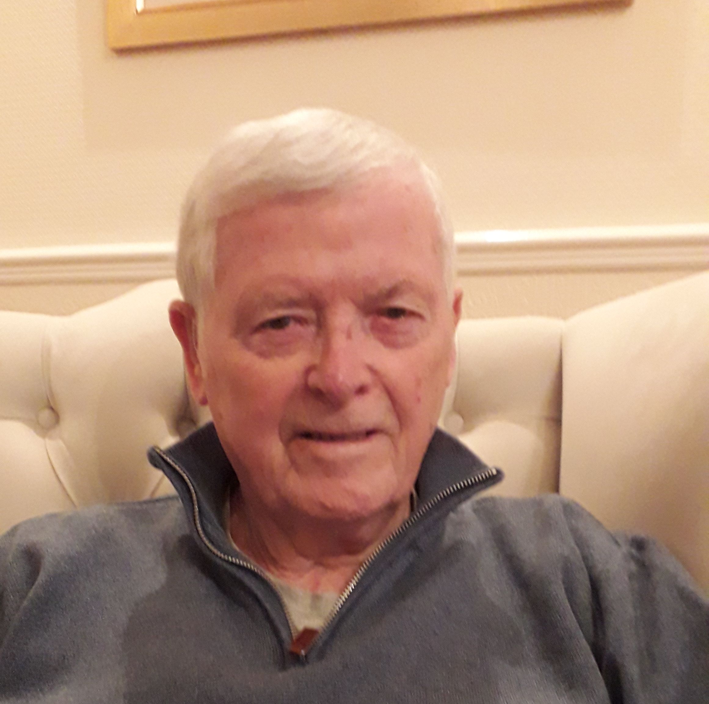 Elderly man with short white hair, smiling gently while seated on a cream upholstered chair, wearing a blue pullover.