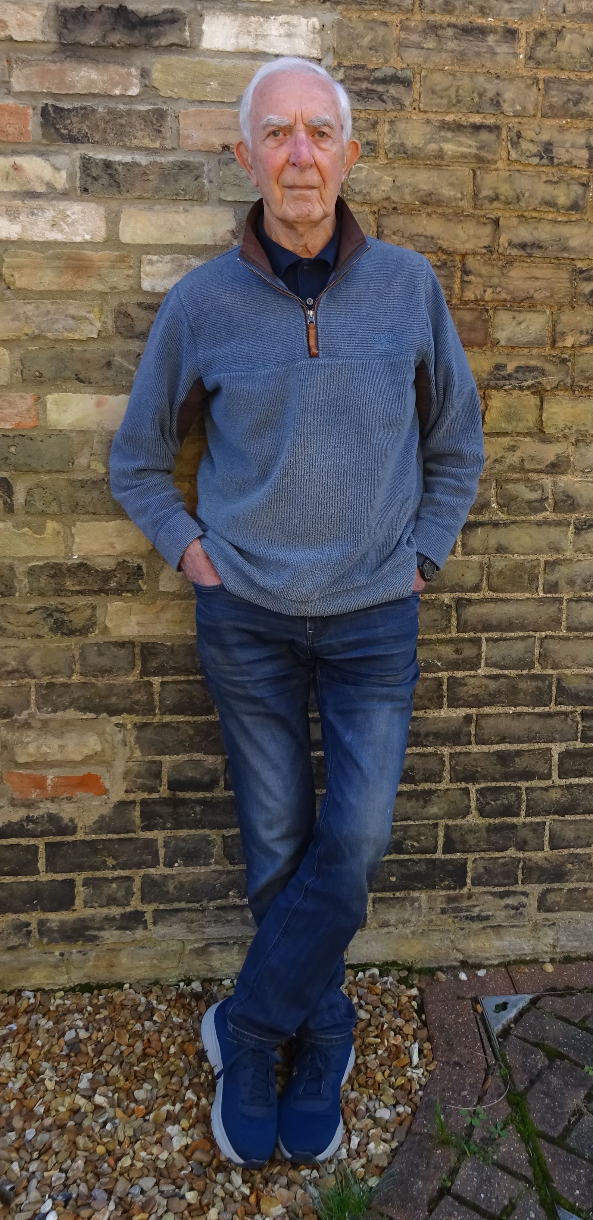 Elderly man with gray hair wearing a blue pullover, dark shirt, jeans, and sneakers, standing against a brick wall with his hands in his pockets and legs crossed.