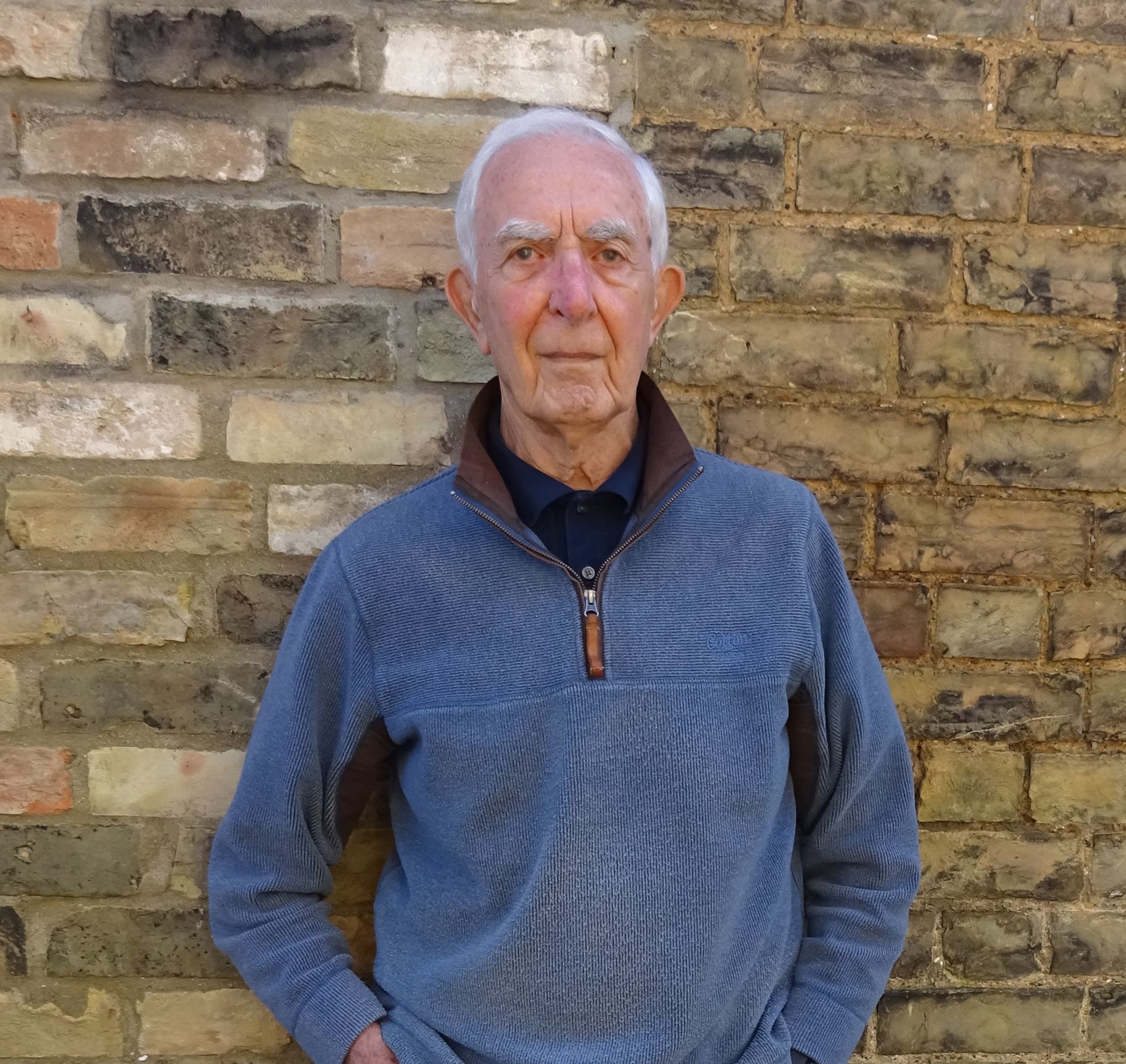 Elderly man with gray hair wearing a blue pullover, dark shirt, jeans, and sneakers, standing against a brick wall with his hands in his pockets and legs crossed.