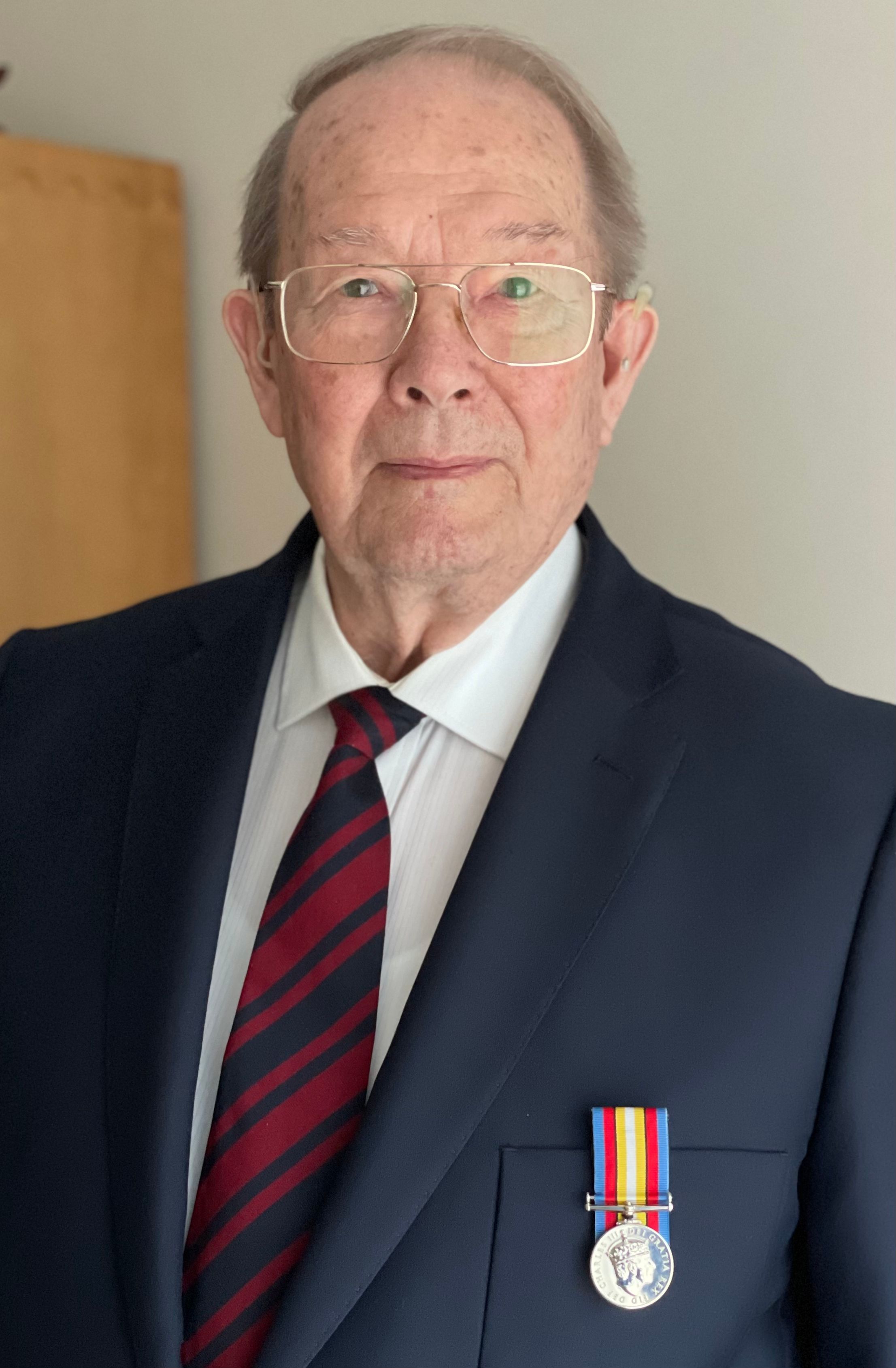Elderly man wearing glasses, a dark suit with a red and black striped tie, and a medal pinned to his jacket, looking directly at the camera with a composed expression.