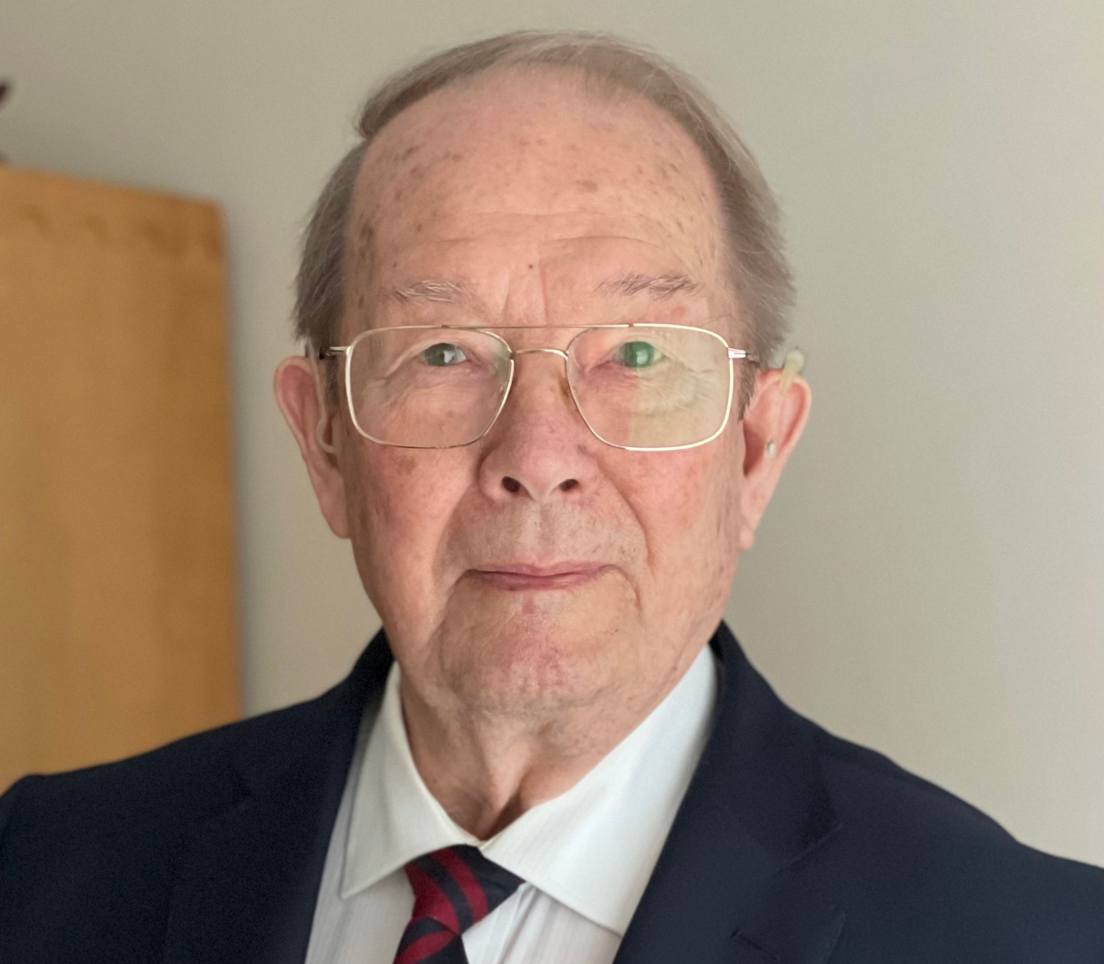Elderly man wearing glasses, a dark suit with a red and black striped tie, and a medal pinned to his jacket, looking directly at the camera with a composed expression.