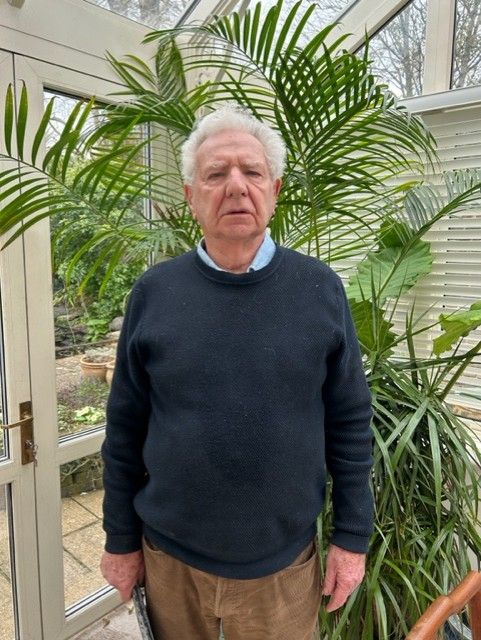 Elderly man with white hair and a serious expression, wearing a dark sweater over a collared shirt and brown trousers, standing in front of tall green plants inside a conservatory.