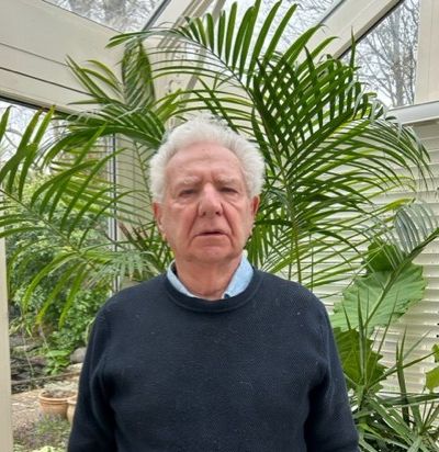 Elderly man with white hair and a serious expression, wearing a dark sweater over a collared shirt and brown trousers, standing in front of tall green plants inside a conservatory.
