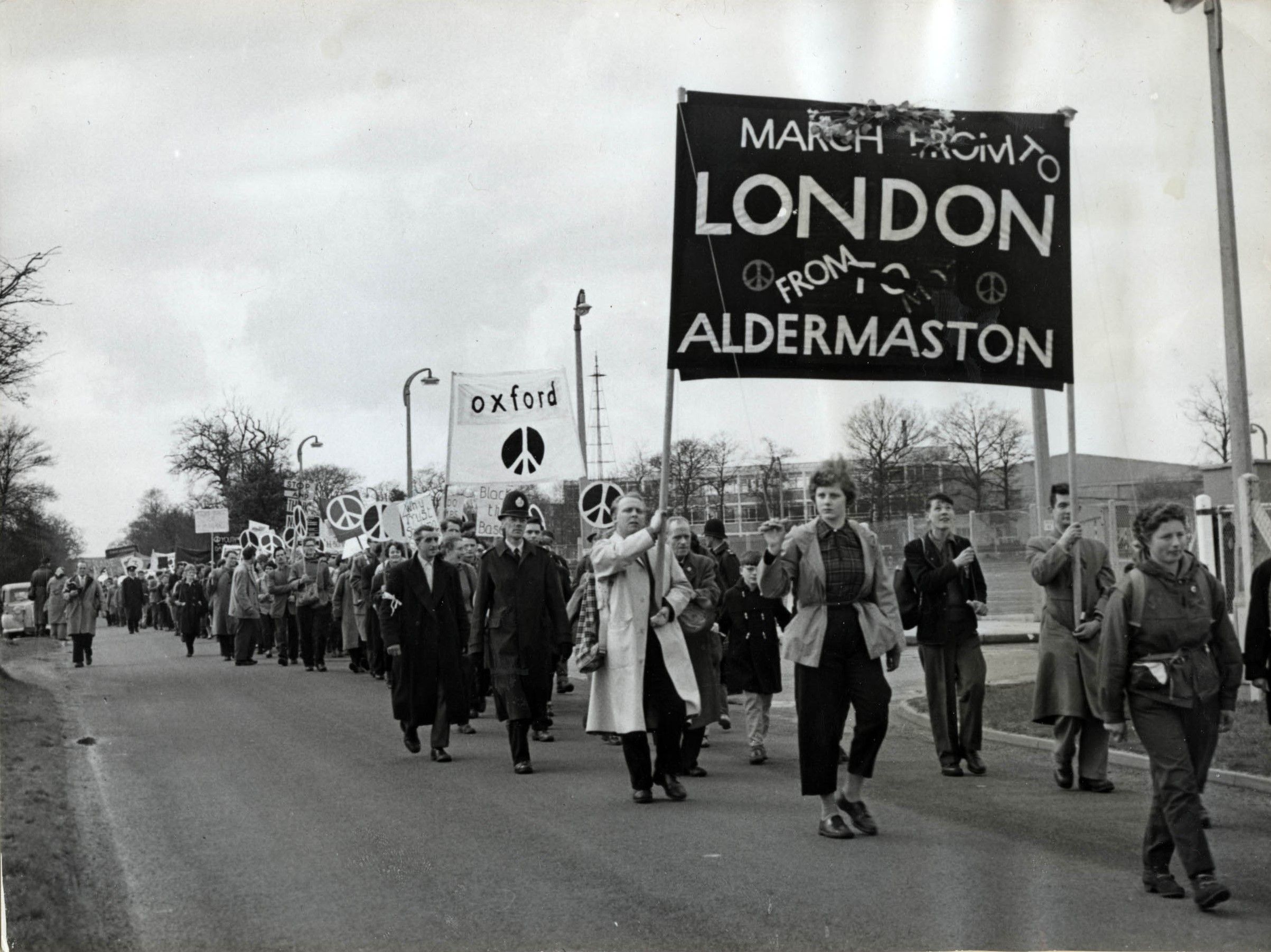 Demonstrators march on the road that passes the chain link fence of the Aldermaston Weapons Research Establishment. At the front of the march, demonstrators hold up a large black banner on poles, which in white text reads: 'From London to Aldermaston'.