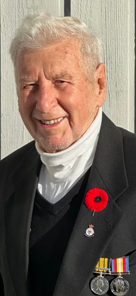 Elderly man smiling, wearing a dark blazer with medals and a red poppy pin, along with a white turtleneck and black sweater underneath.