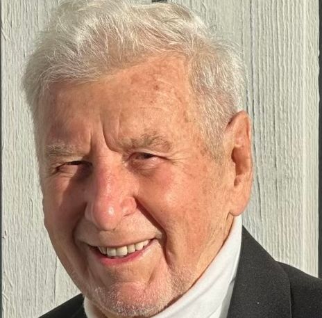 Elderly man smiling, wearing a dark blazer with medals and a red poppy pin, along with a white turtleneck and black sweater underneath.