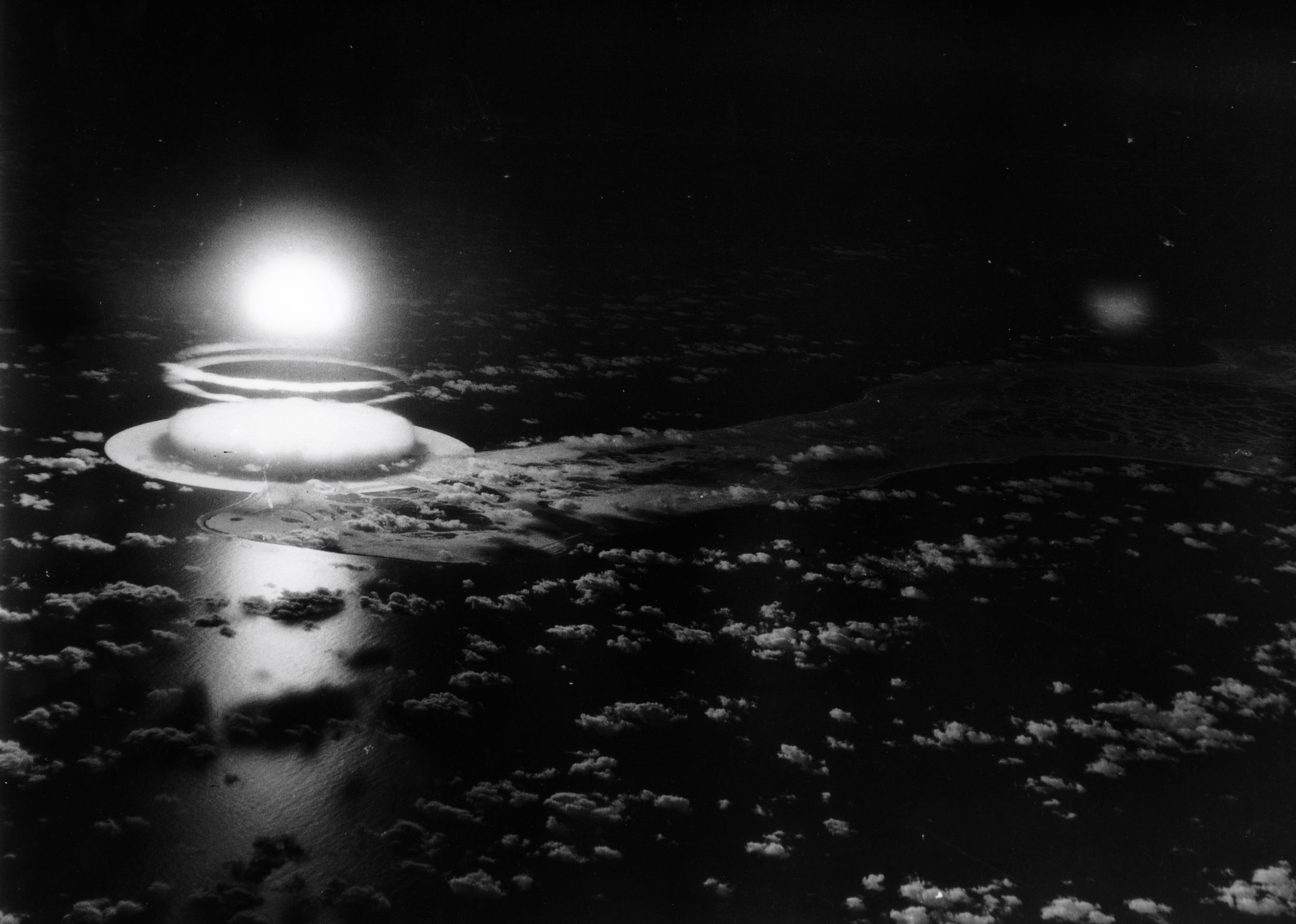 A black-and-white image of the far-east point of Christmas Island, which is illuminated by the brilliant blast of Halliard, itself forming a glowing bulb over the Pacific Ocean.
