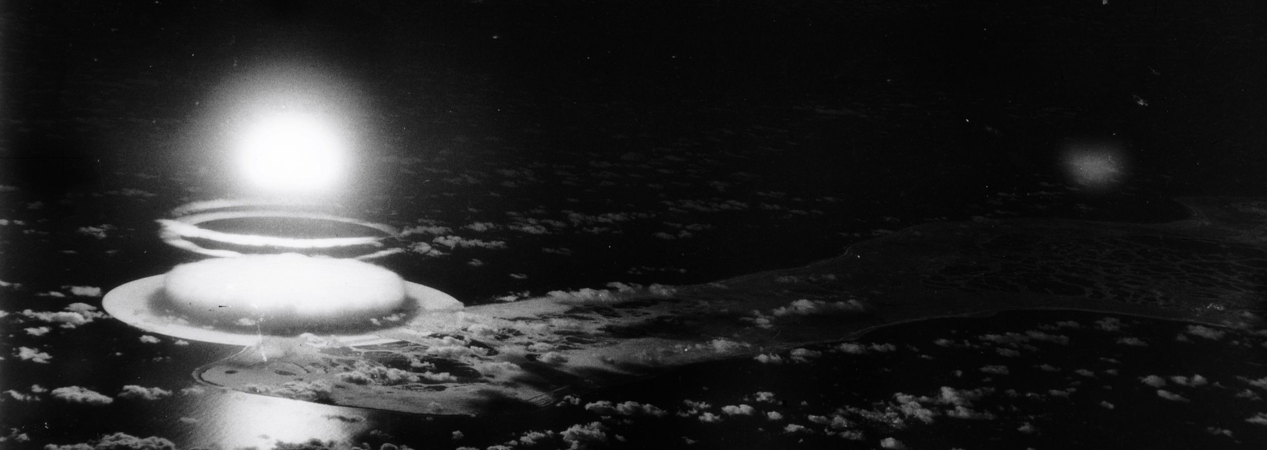 A black-and-white image of the far-east point of Christmas Island, which is illuminated by the brilliant blast of Halliard, itself forming a glowing bulb over the Pacific Ocean.