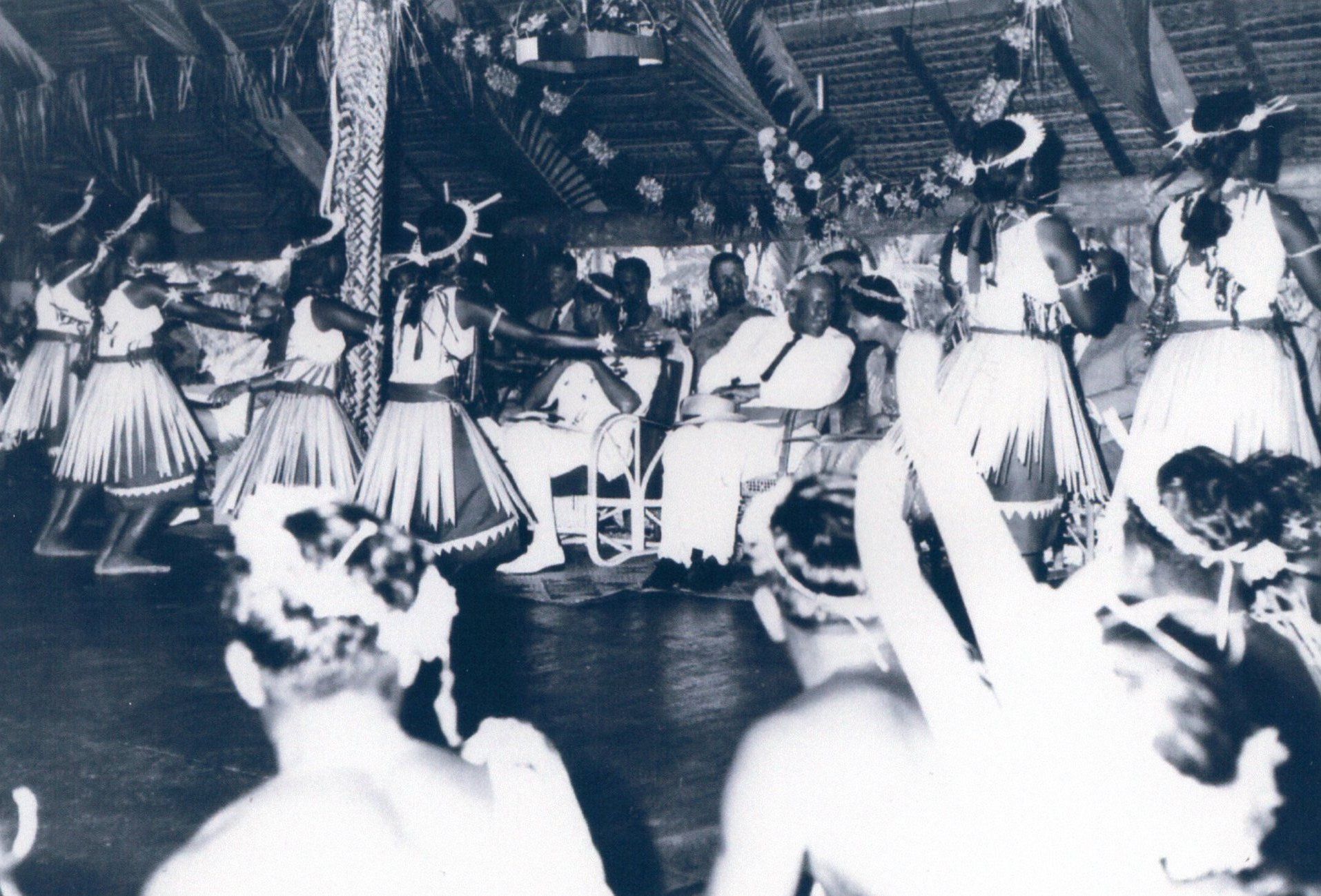 Young I-Kiribati women with grass skirts and headdresses perform a dance in front of dignitaries wearing white in a maneaba, including Prince Philip.