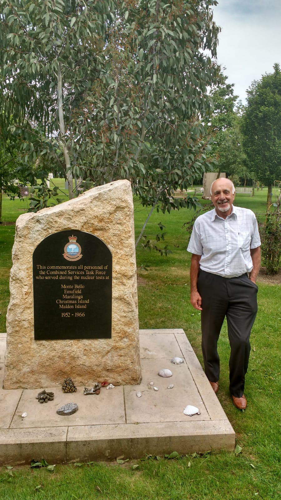 Elderly man smiling with hands in his pockets, wearing a short-sleeved checked shirt and dark trousers, standing beside a large stone memorial plaque set in a grassy park with trees in the background.