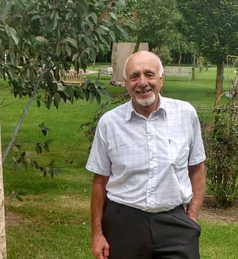 Elderly man smiling with hands in his pockets, wearing a short-sleeved checked shirt and dark trousers, standing beside a large stone memorial plaque set in a grassy park with trees in the background.