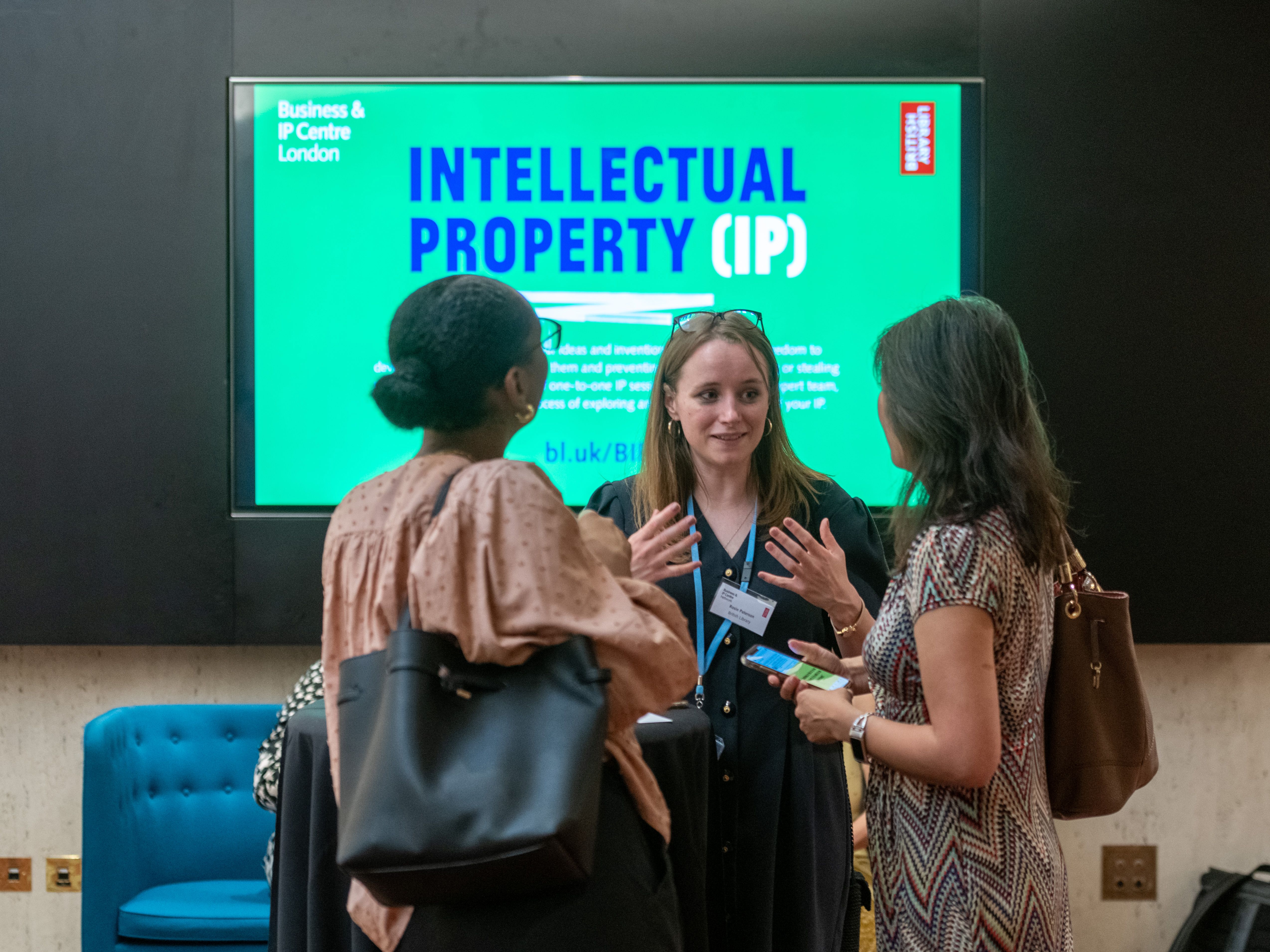 Three women talking at the BIPC with Intellectual Property digital display in background.