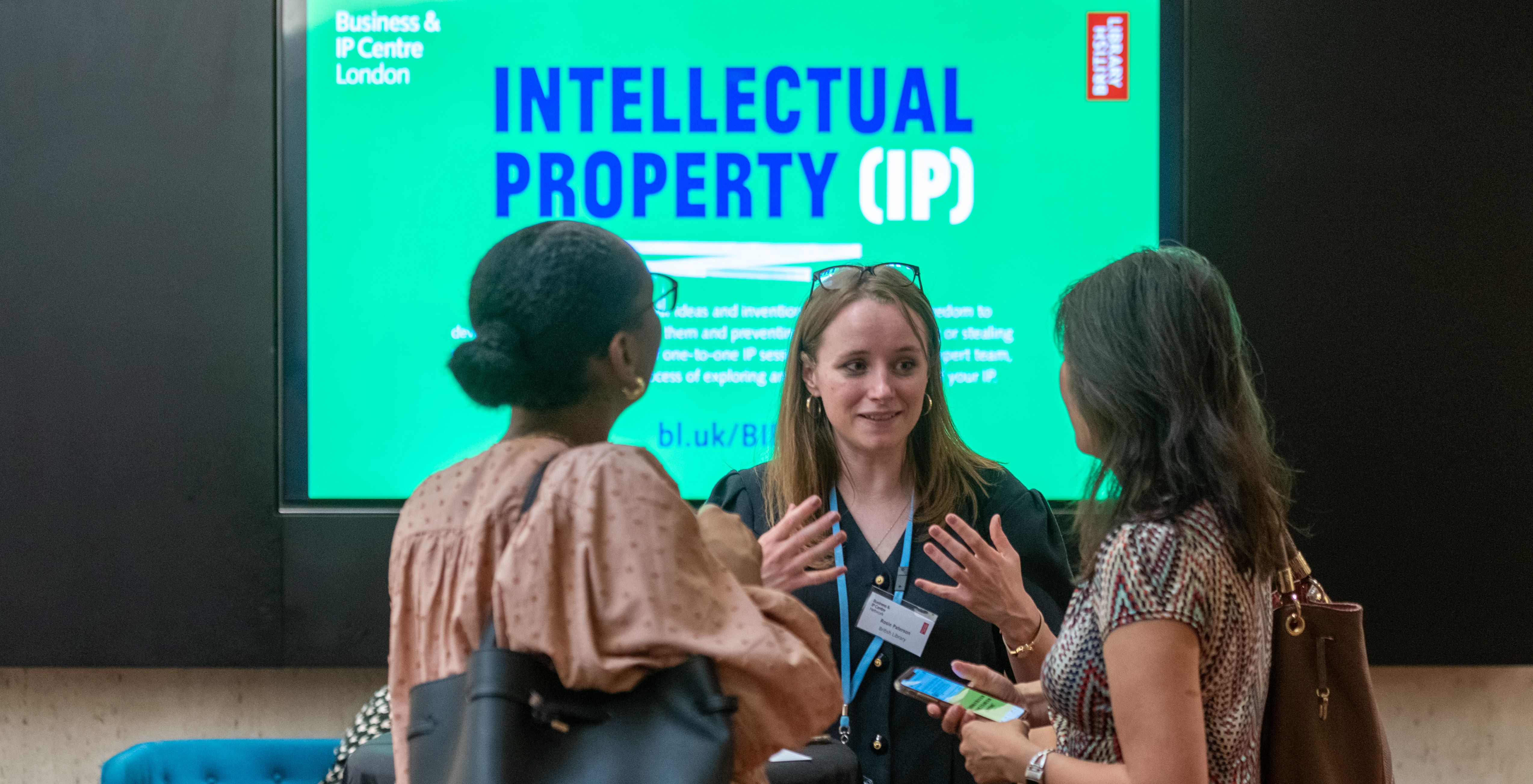 Three women talking at the BIPC with Intellectual Property digital display in background.