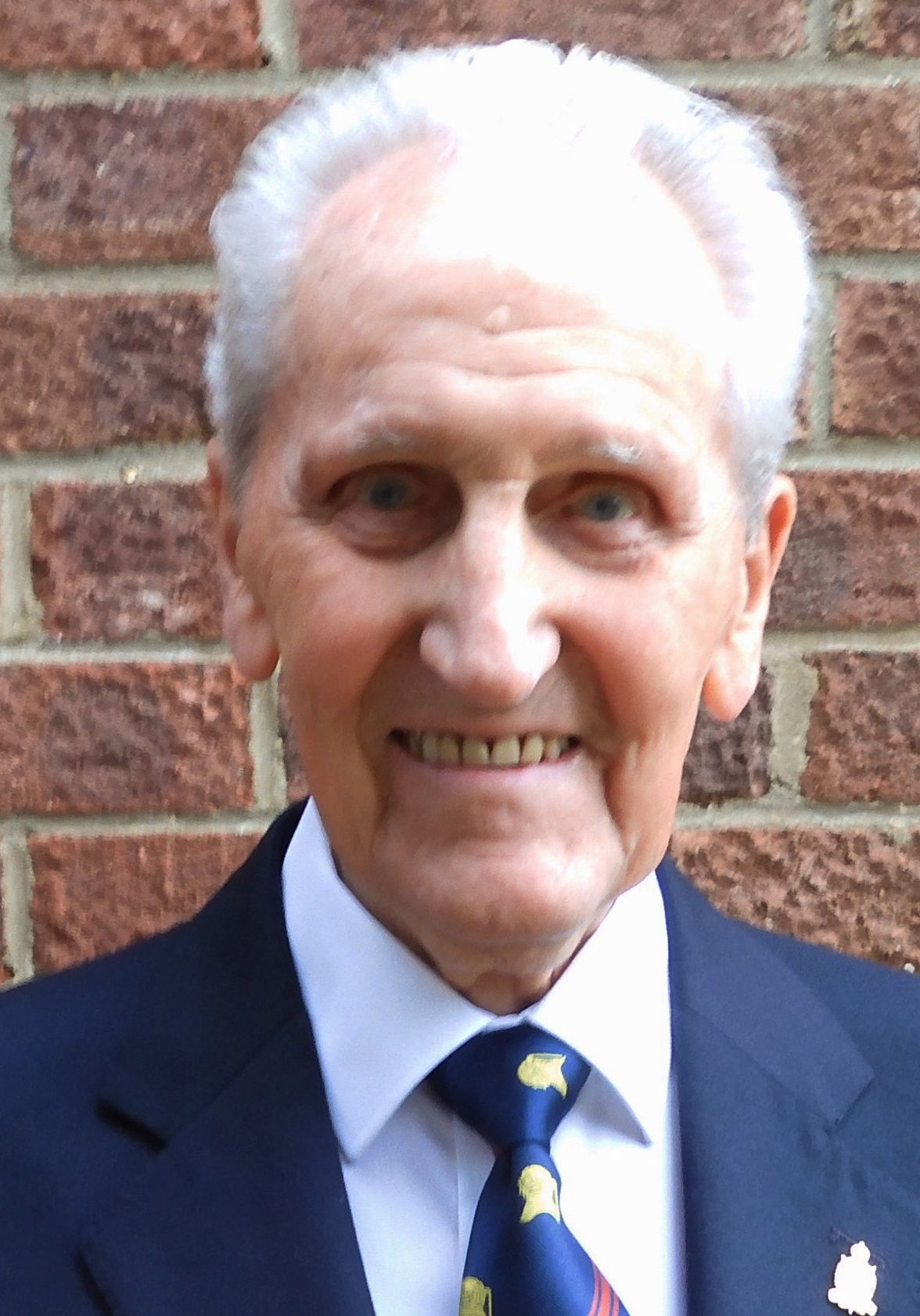 Elderly man smiling, wearing a dark suit jacket with a small lapel pin, white shirt, and patterned tie, standing in front of a brick wall.