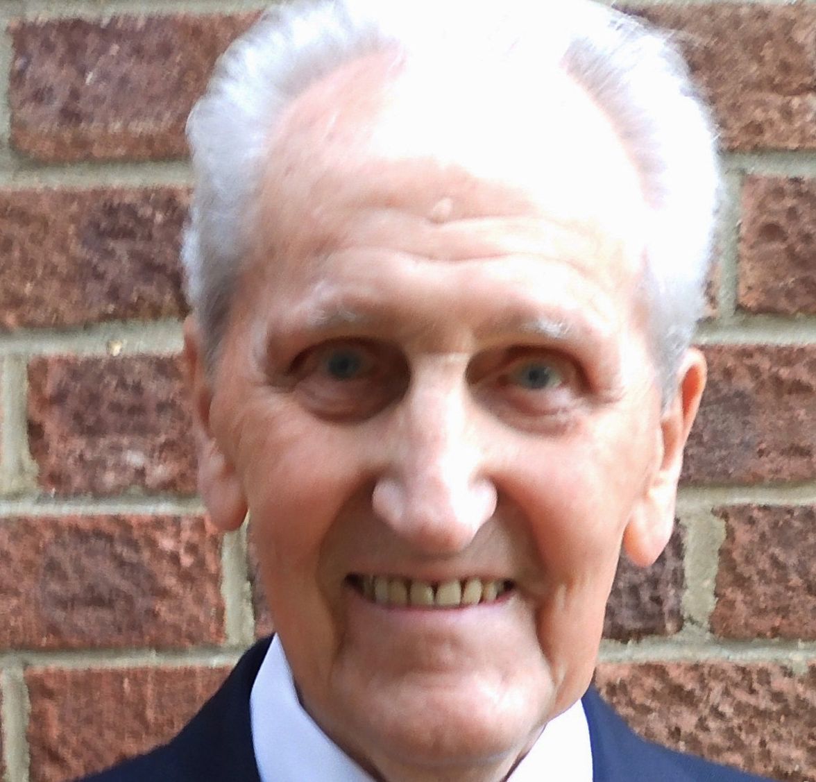 Elderly man smiling, wearing a dark suit jacket with a small lapel pin, white shirt, and patterned tie, standing in front of a brick wall.