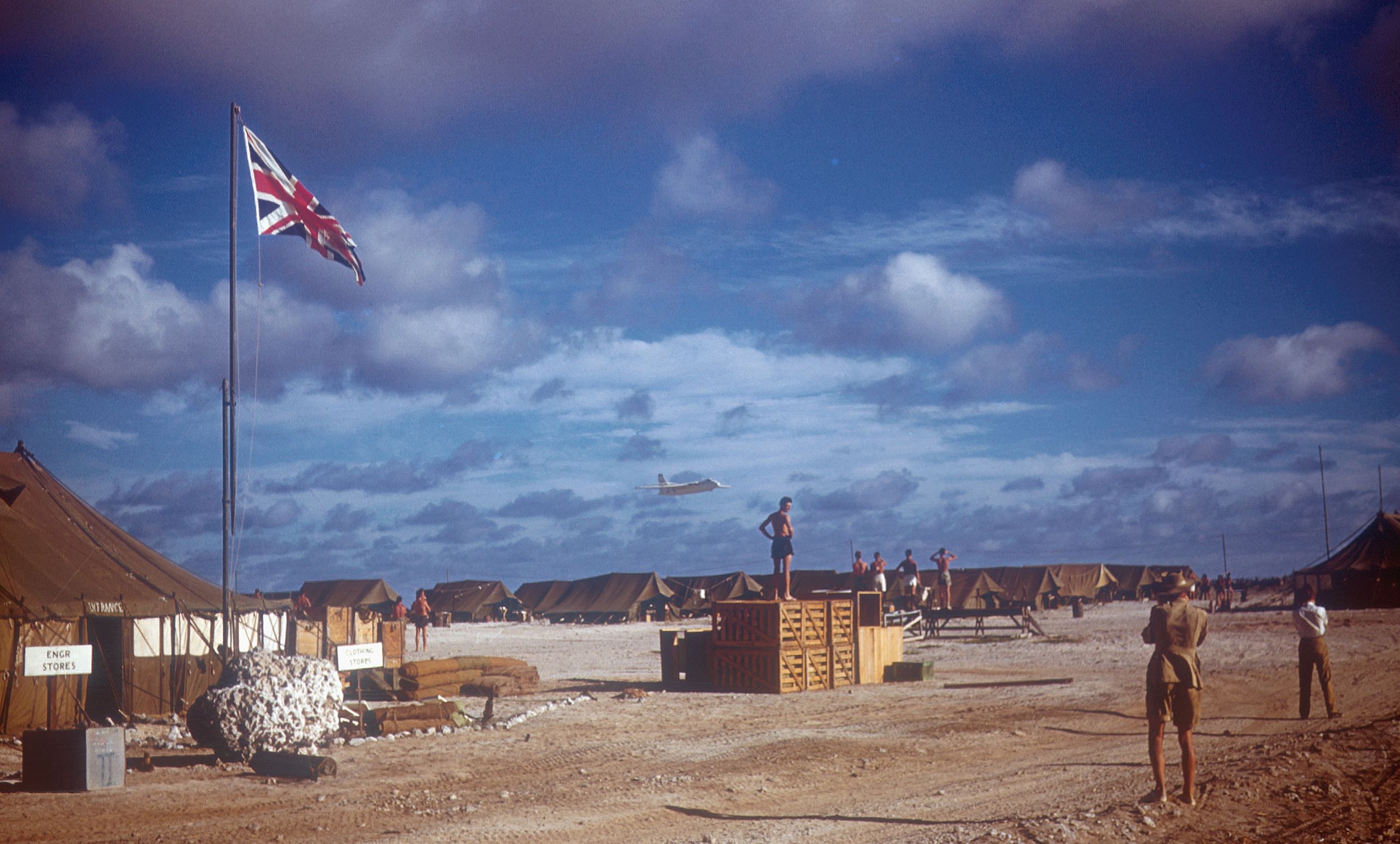Khaki-coloured tents line the background. A British flag is waving in the air, having been fully hoisted on the mast. Soldiers are chatting and walking around the camp.