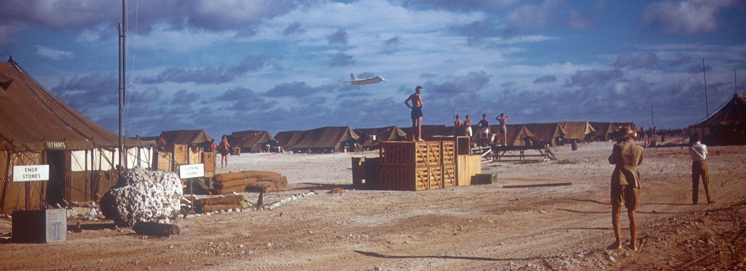 Khaki-coloured tents line the background. A British flag is waving in the air, having been fully hoisted on the mast. Soldiers are chatting and walking around the camp.