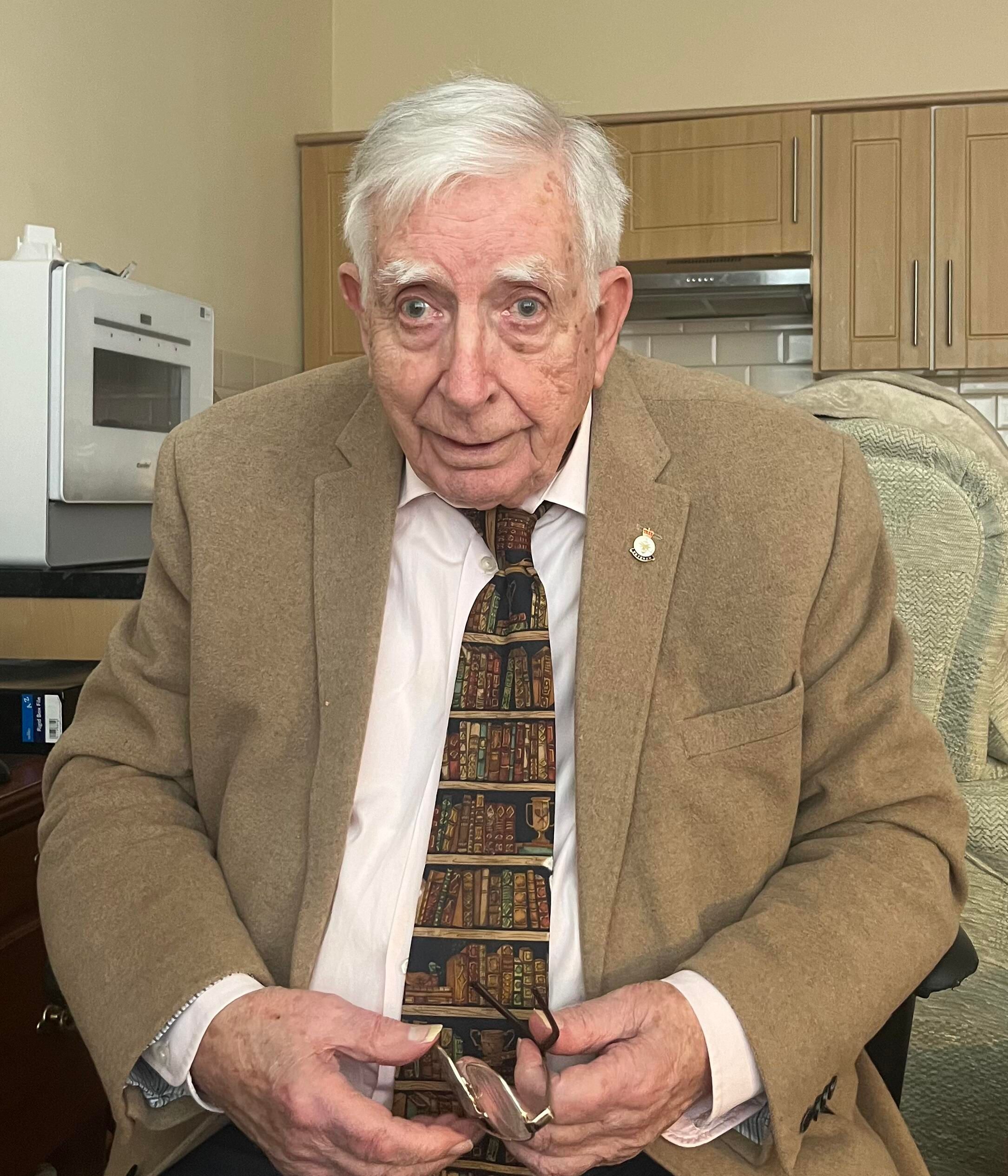 Elderly man seated indoors, wearing a tan blazer, white shirt, and patterned tie decorated with book spines. He holds a pair of glasses in his hands and looks directly at the camera with a calm expression.