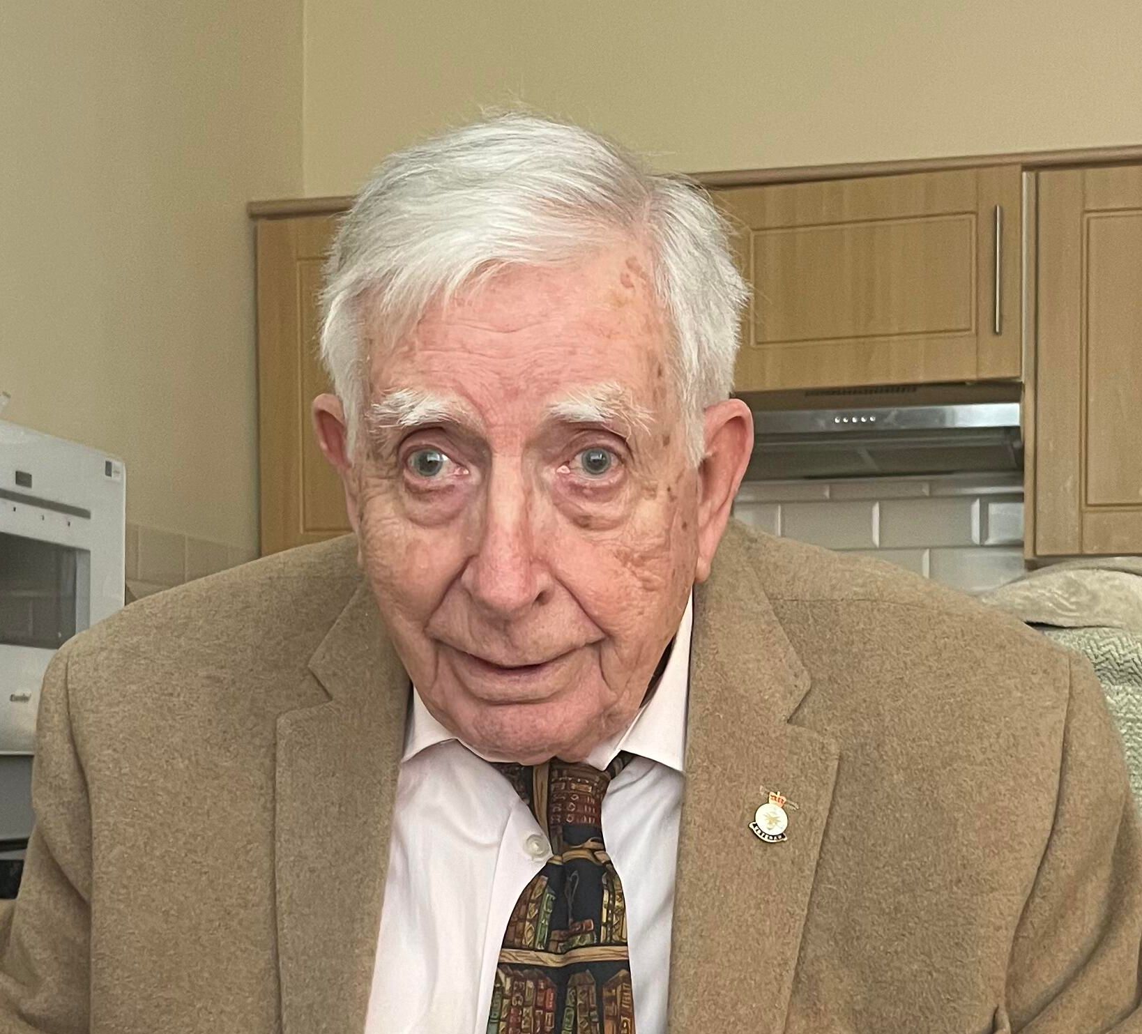 Elderly man seated indoors, wearing a tan blazer, white shirt, and patterned tie decorated with book spines. He holds a pair of glasses in his hands and looks directly at the camera with a calm expression.