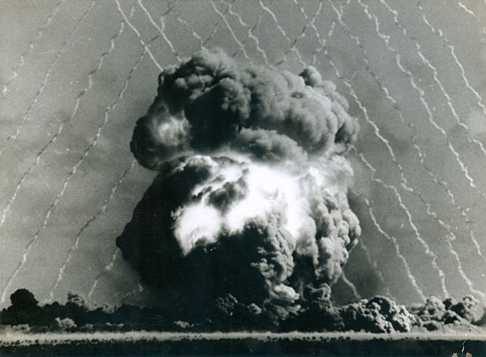 A black-and-white mushroom cloud with rocket trails that criss-cross against the background of the sky.