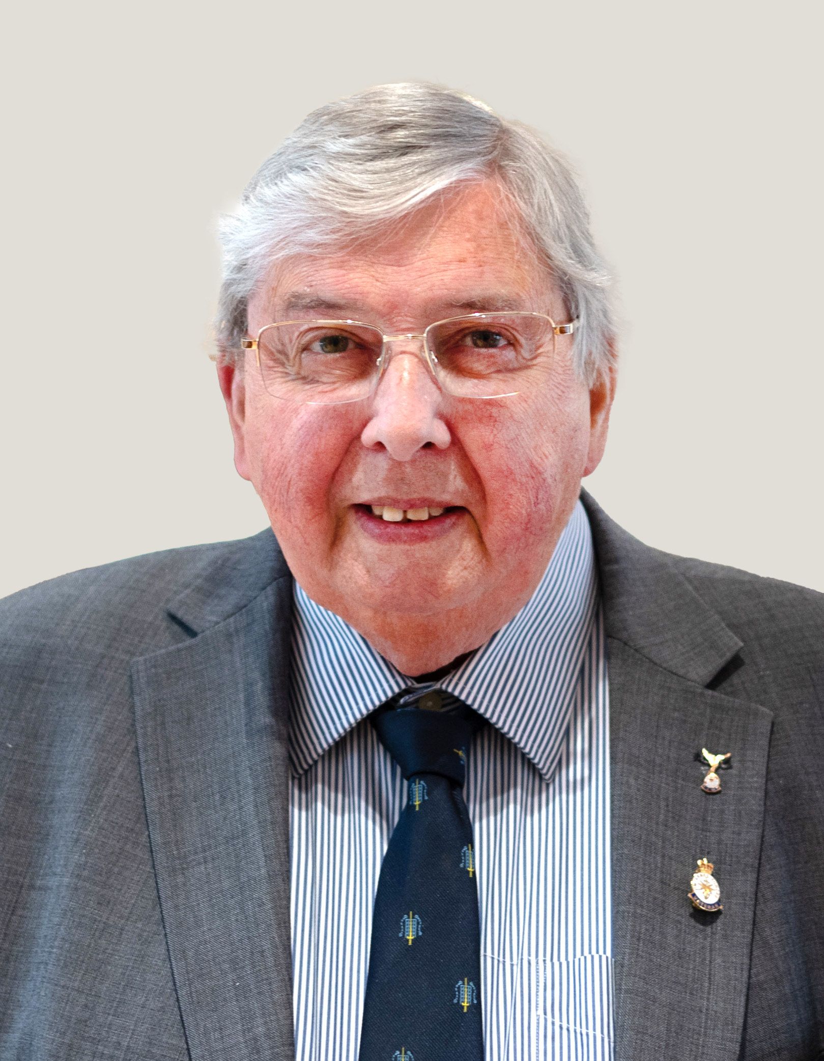 Elderly man smiling, wearing glasses, a gray suit with a striped shirt, dark tie, and two pins on his lapel.