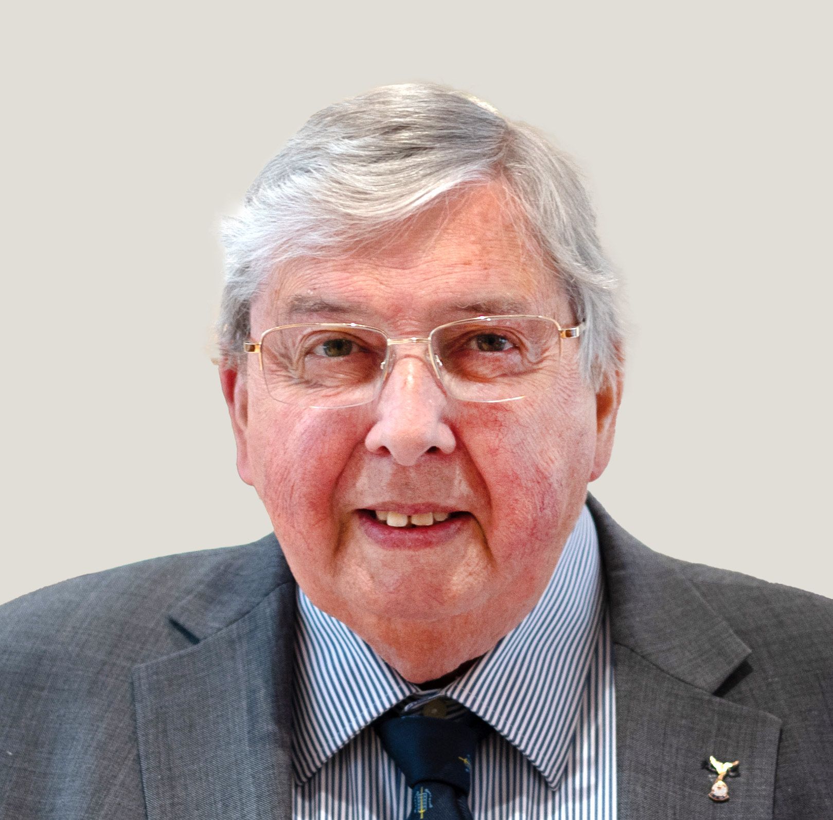 Elderly man smiling, wearing glasses, a gray suit with a striped shirt, dark tie, and two pins on his lapel.