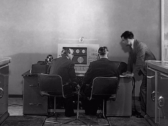 Black-and-white photograph of three men in suits at the console of an early computer. Two are seated with their backs to the camera, while the third stands to the right, leaning toward them. The console in front of them has large dials, switches, and circular displays.