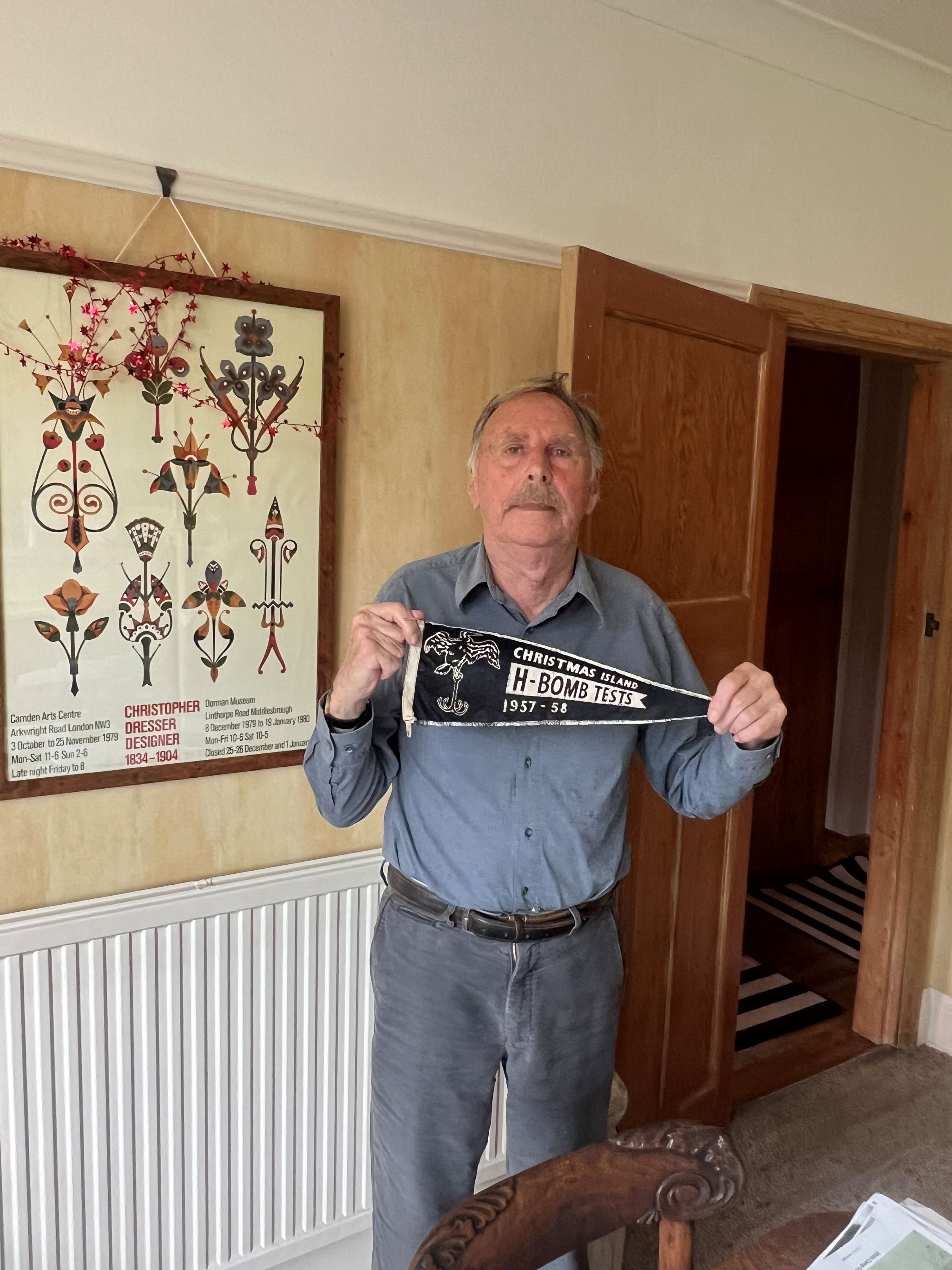 Elderly man with a mustache, wearing a blue shirt and gray trousers, standing and holding a black pennant that reads 'Christmas Island H-Bomb Tests 1957–58.' A framed poster hangs on the wall behind him.