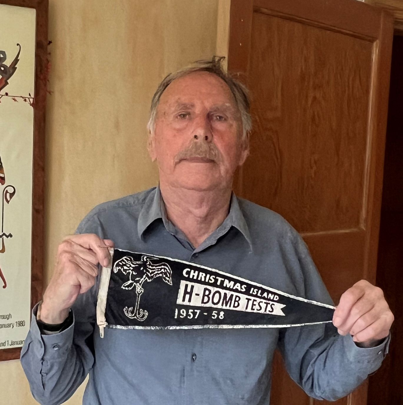 Elderly man with a mustache, wearing a blue shirt and gray trousers, standing and holding a black pennant that reads 'Christmas Island H-Bomb Tests 1957–58.' A framed poster hangs on the wall behind him.