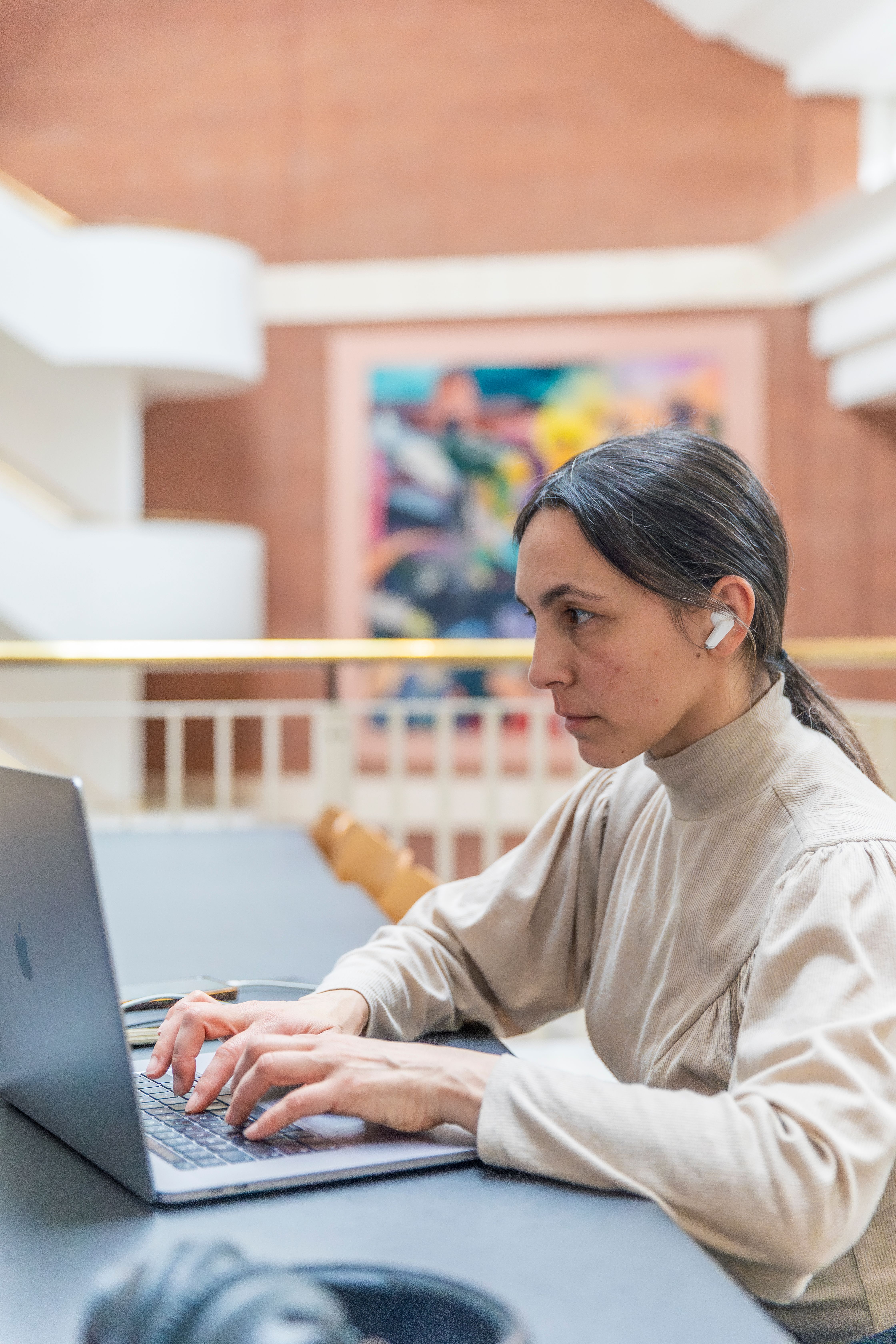 A woman sits, looking serious and wearing in-ear headphones, at a laptop, in the public study spaces of the British Library in London.