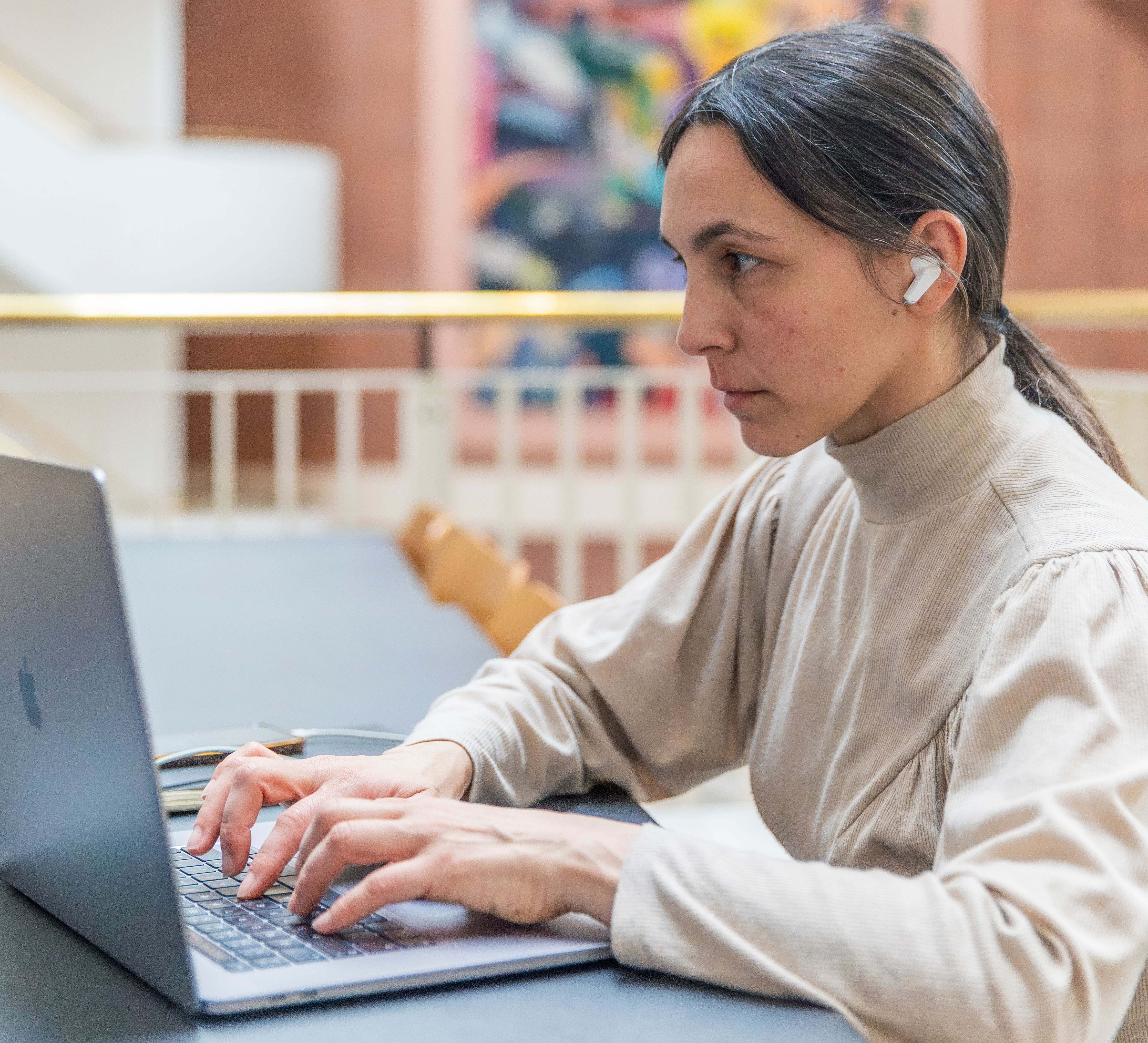 A woman sits, looking serious and wearing in-ear headphones, at a laptop, in the public study spaces of the British Library in London.