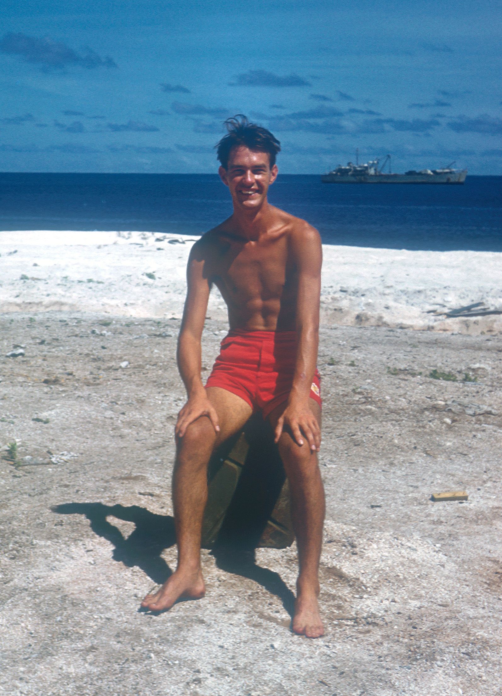 Young man smiling and sitting on a rock or crate, shirtless and wearing red shorts, on a sandy beach with the ocean and a ship in the background.