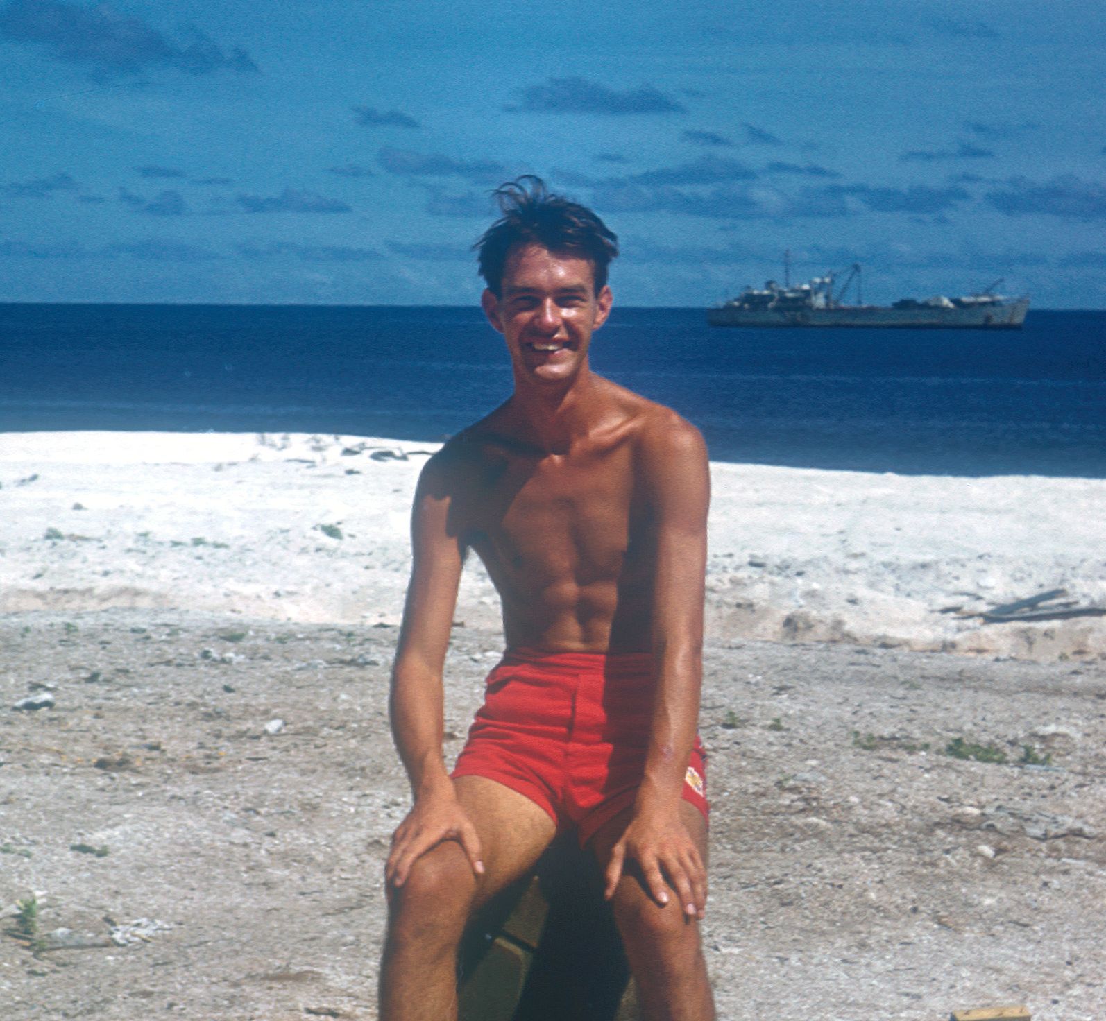 Young man smiling and sitting on a rock or crate, shirtless and wearing red shorts, on a sandy beach with the ocean and a ship in the background.