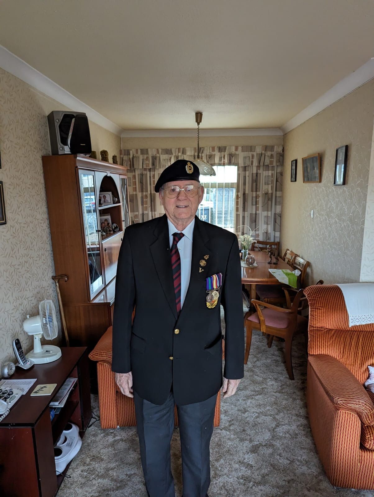 Elderly man wearing glasses, a beret with an insignia, a dark blazer with medals, and a striped tie, standing in a living room with orange armchairs and a cabinet behind him.
