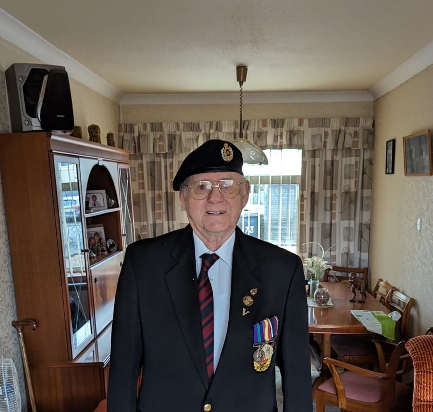 Elderly man wearing glasses, a beret with an insignia, a dark blazer with medals, and a striped tie, standing in a living room with orange armchairs and a cabinet behind him.